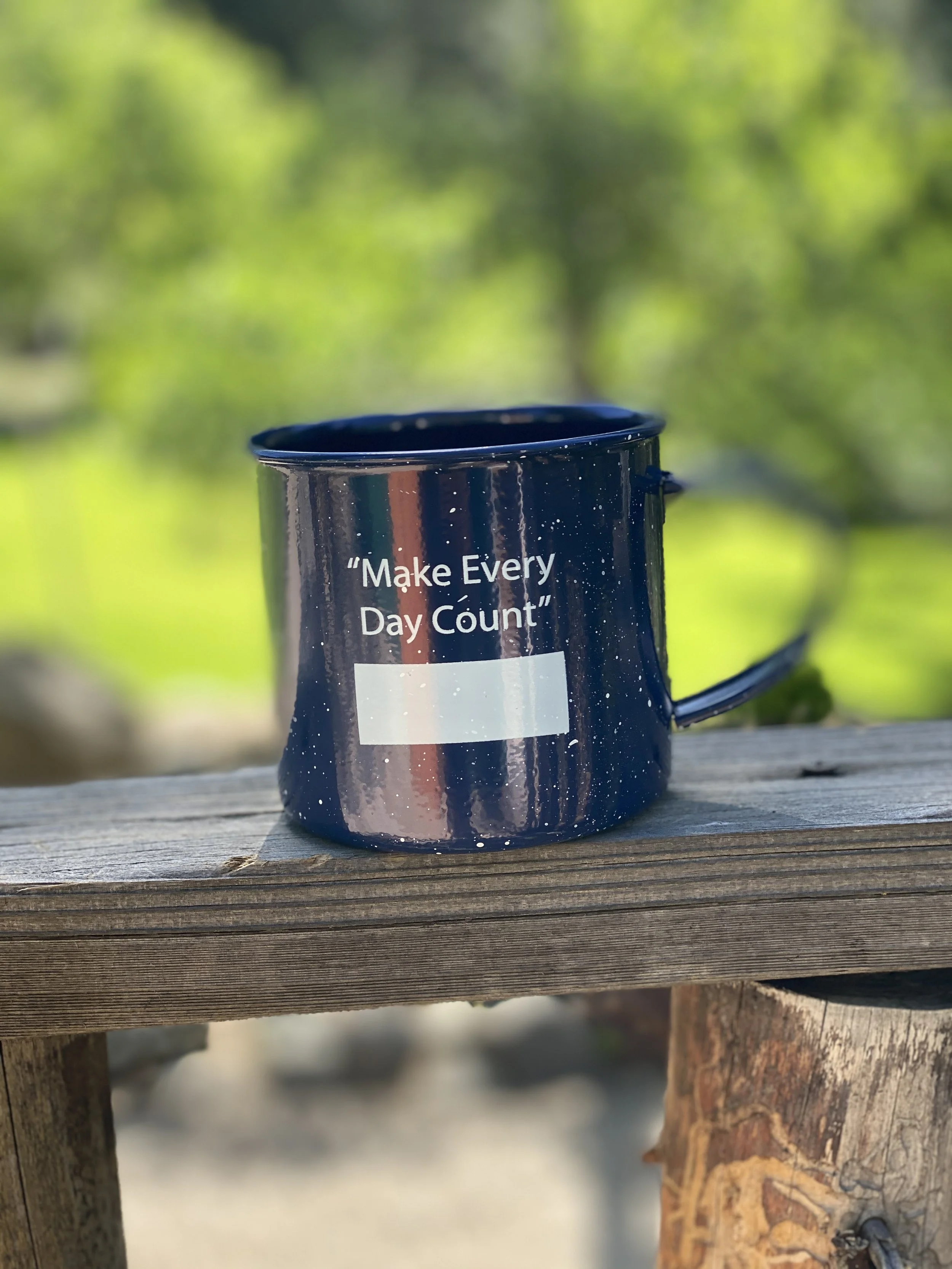 Black mug with white text that says 'Make Every Day Count' perched on a weathered wooden outdoor table, with a blurred green natural background.