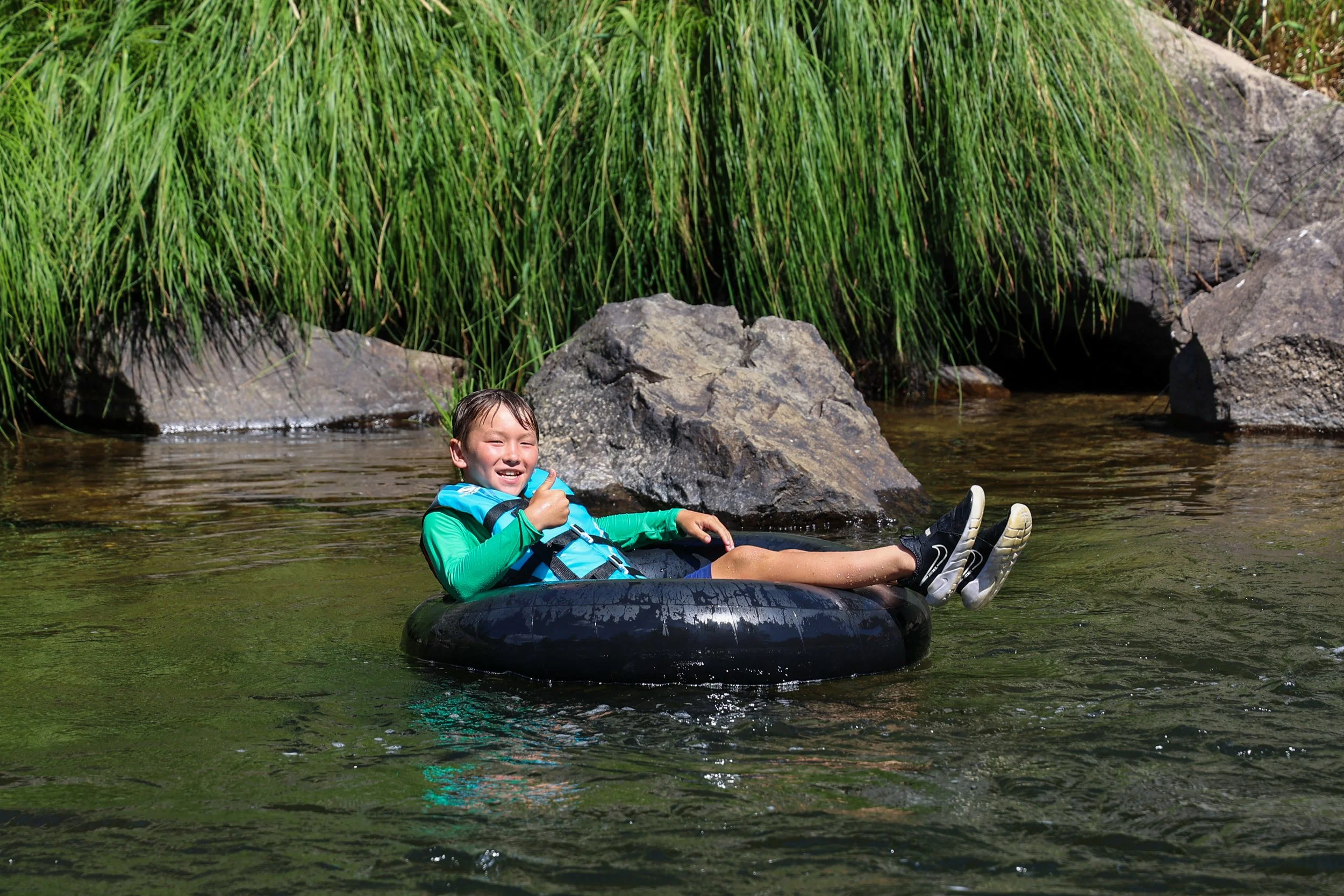 Child floating on a black inner tube in a river, giving a thumbs-up, surrounded by rocks and green tall grass.