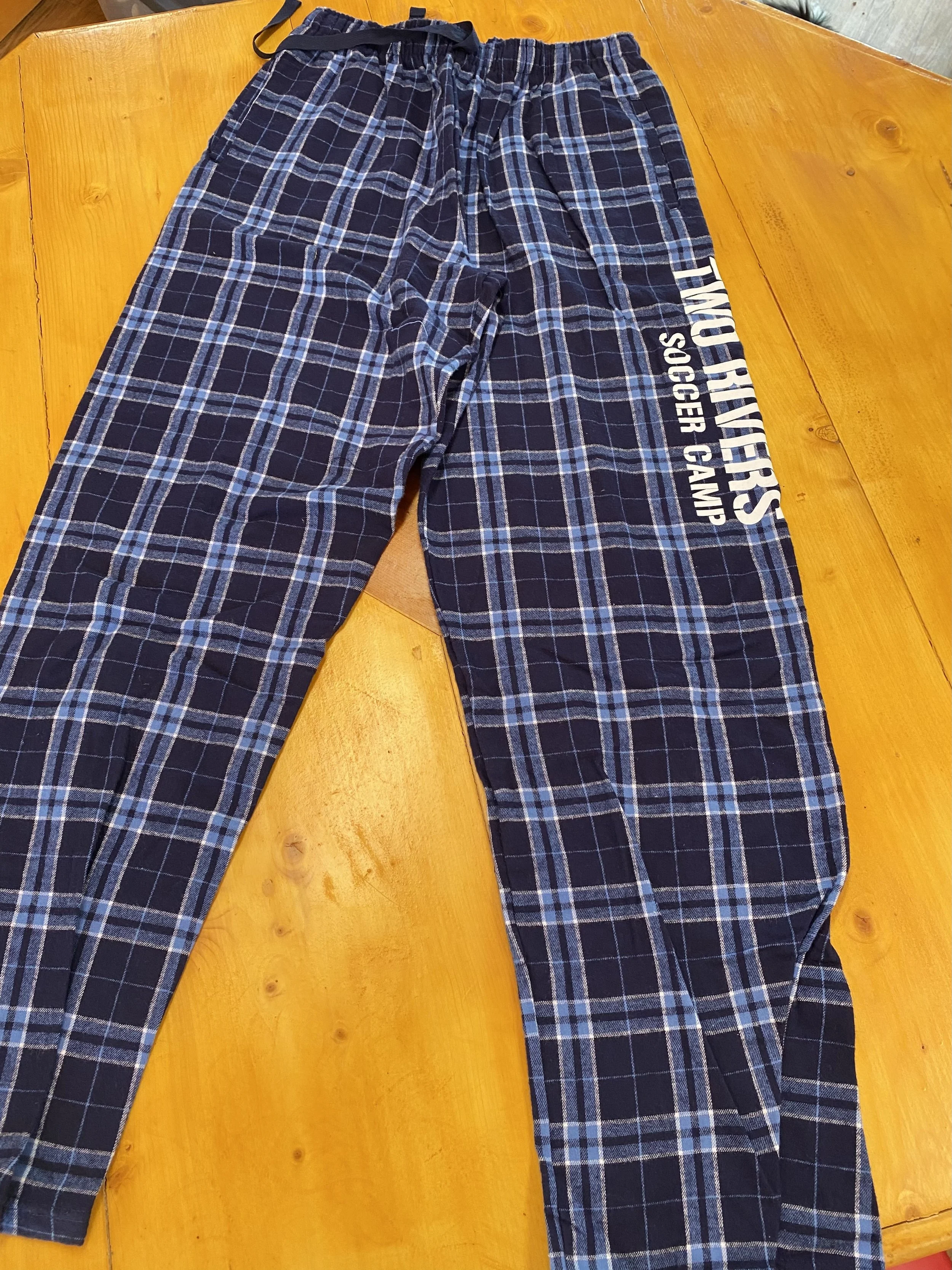 Blue plaid pajama pants with white text on the right thigh that reads 'TWO RIVERS SOCCER CAMP,' laid flat on a wooden surface.