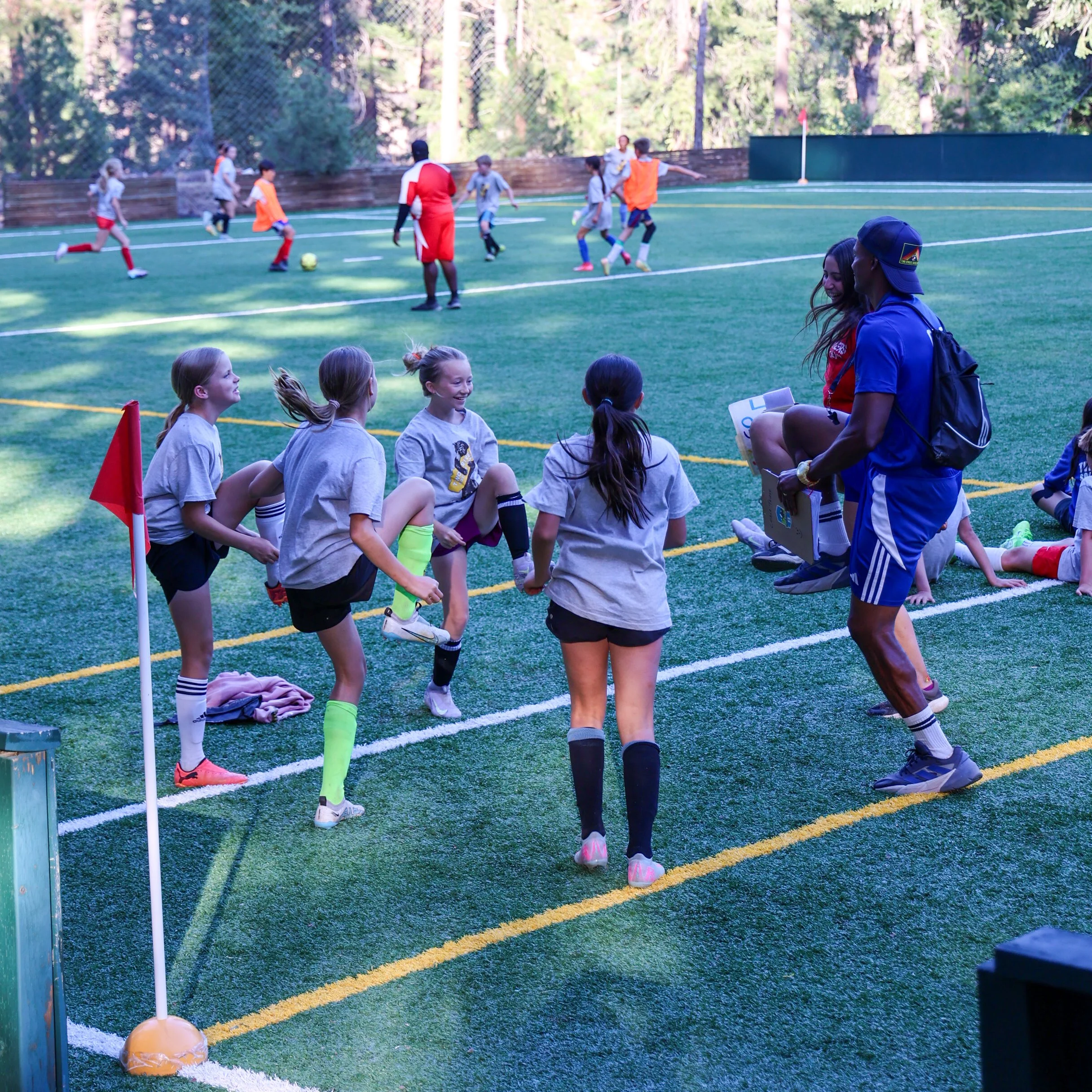 Youth soccer practice on a turf field with kids and coaches, some sitting on the sideline and others playing in the background at a wooded park.