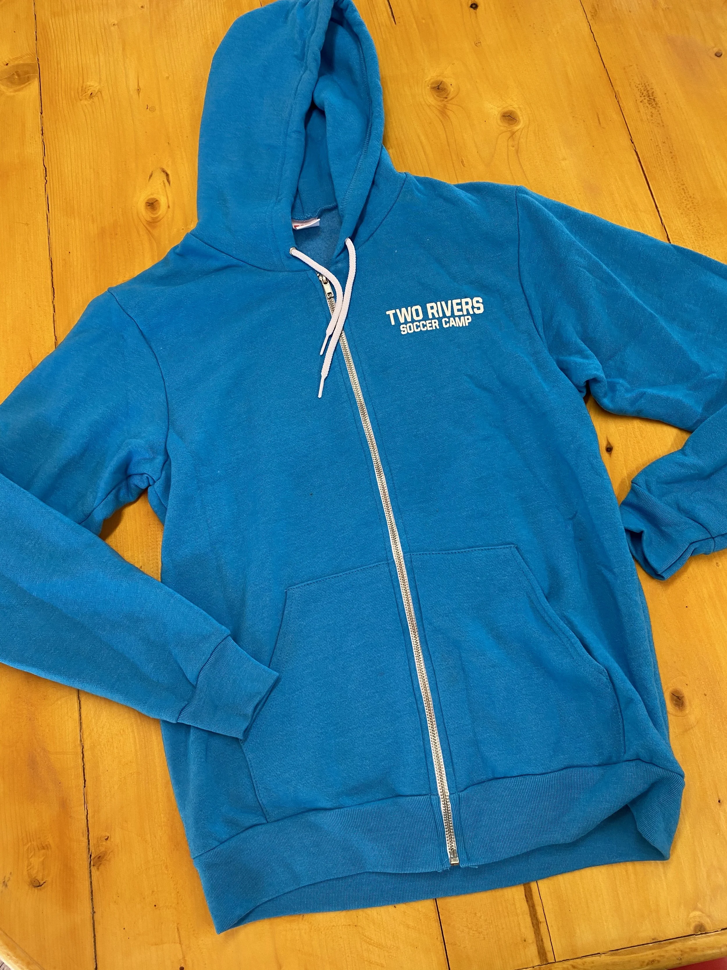 Blue full-zip hoodie with 'Two Rivers Soccer Camp' text on the chest, placed on a wooden surface.