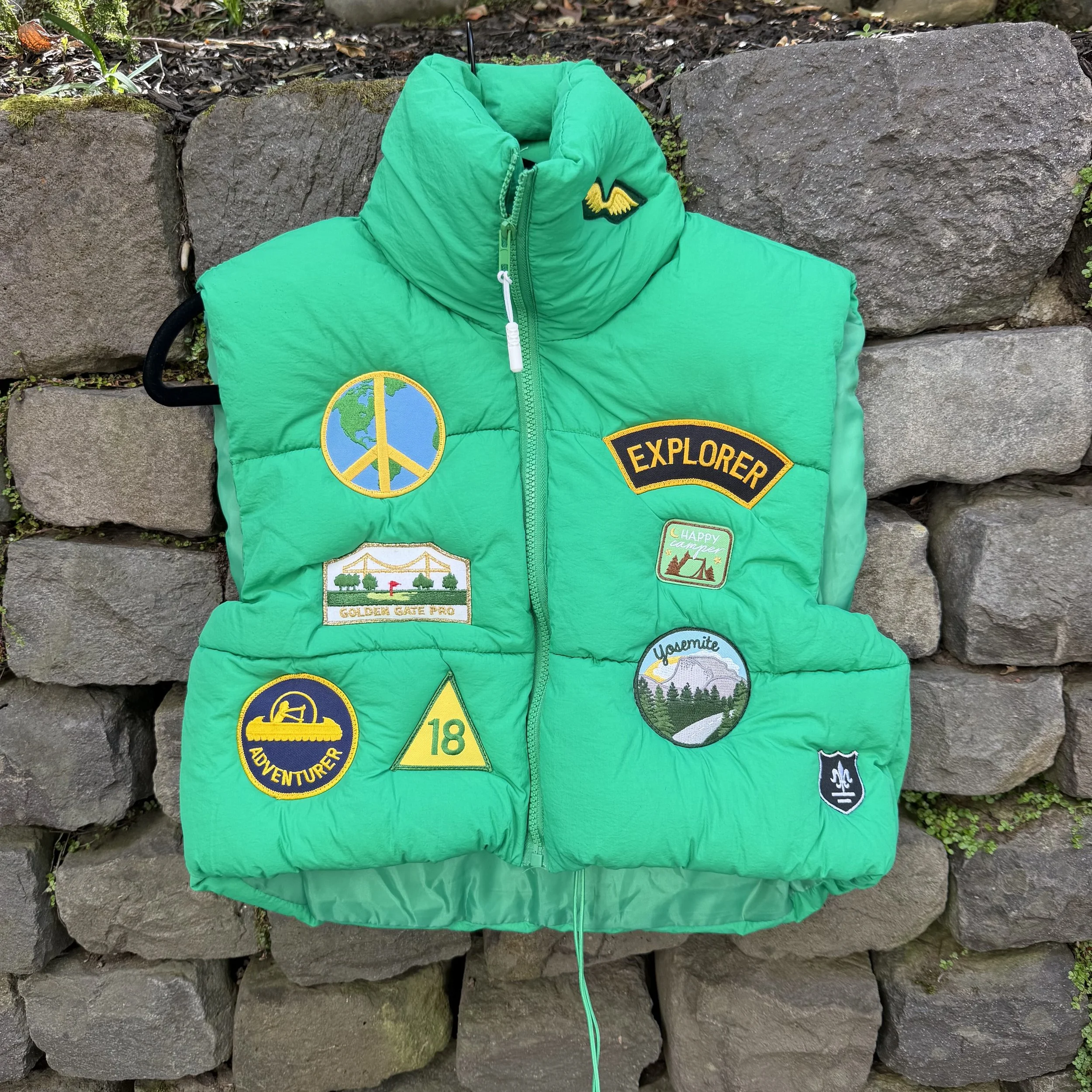 A green puffer vest with various adventure-themed patches, including a peace sign with Earth, a yellow 'EXPLORER' patch, a camper van with 'HAPPY CAMPER,' a mountain and forest scene with 'Yosemite,' a yellow triangle with '18,' and a black and blue 