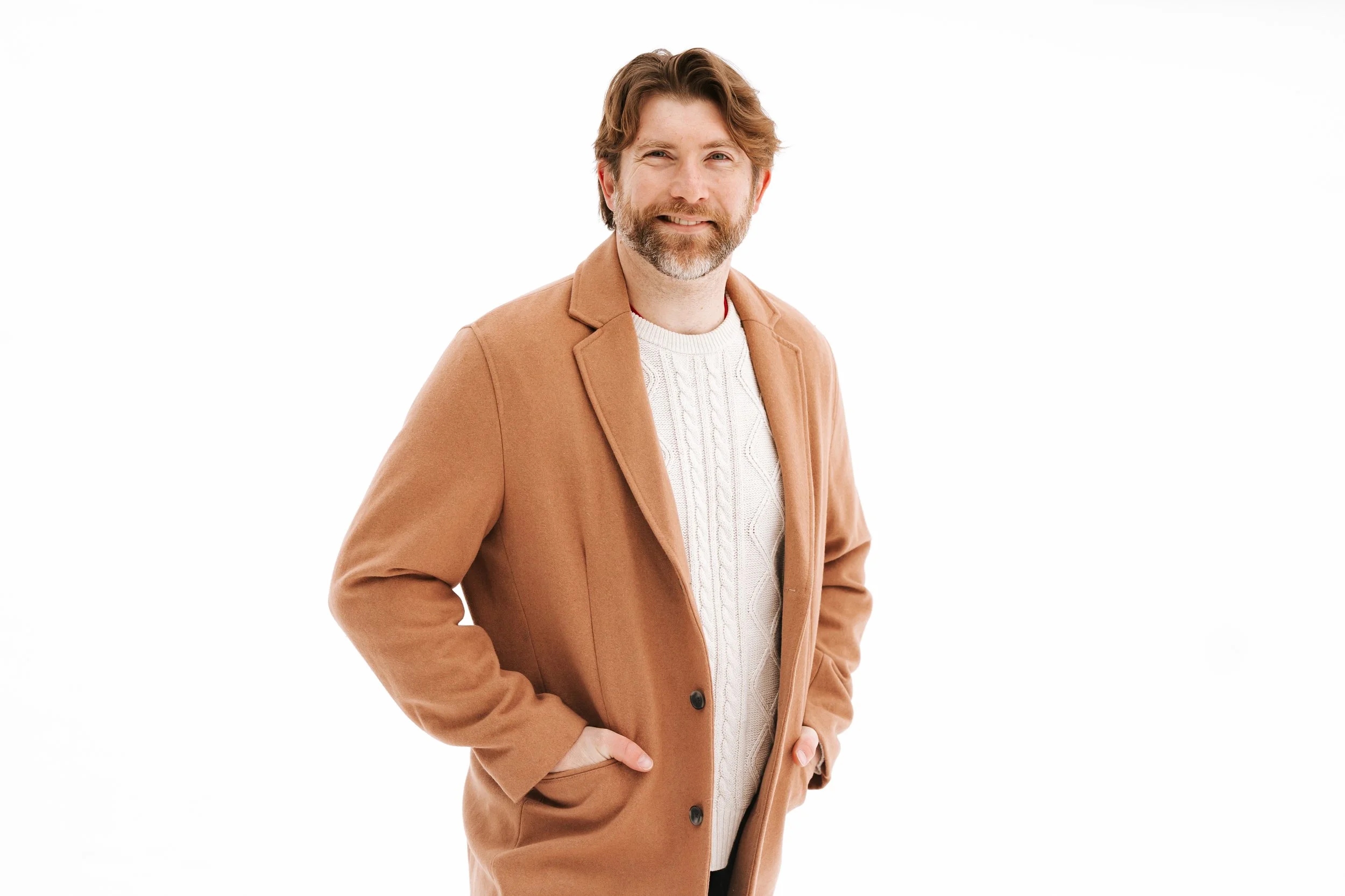 Man with brown hair and beard smiling, wearing a tan coat and a white cable knit sweater, standing with his hands in his pockets against a white background.