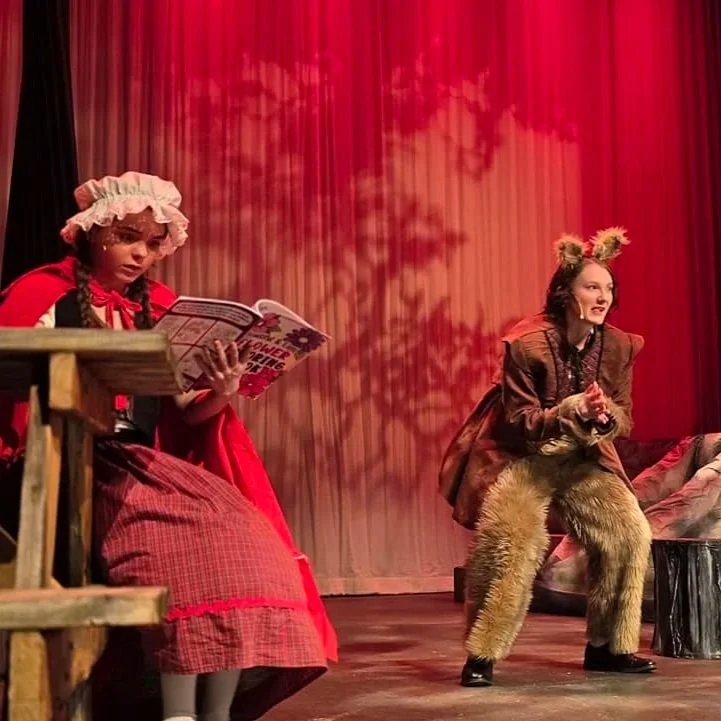 Children in costume performing on stage with red curtains. One girl is dressed as Little Red Riding Hood reading a book, and another girl is dressed as a wolf or animal character with furry pants and animal ears, appearing to be speaking or singing.