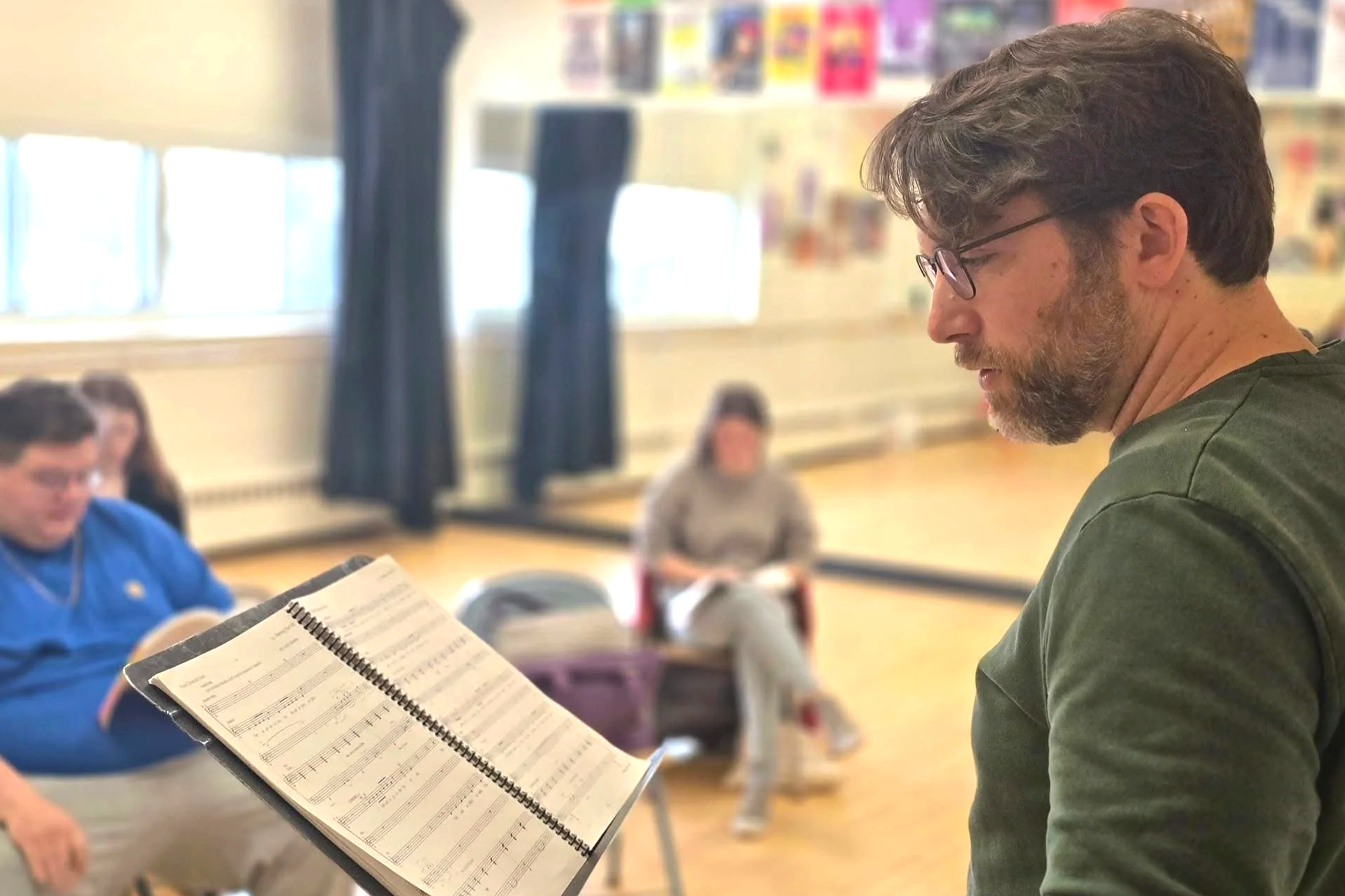 A man with glasses and a beard standing in a classroom, reading from a music book or sheet music. In the background, three people are seated and appear to be listening or watching.