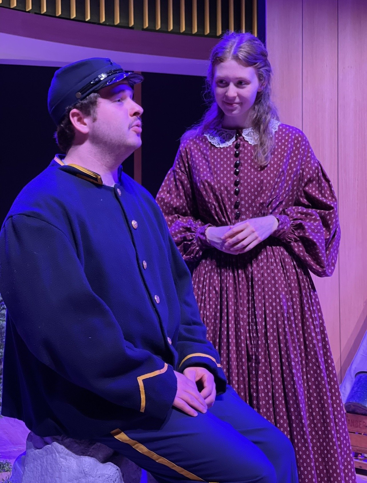 Two actors in period costumes, one sitting and the other standing, during a scene in a theatrical production.