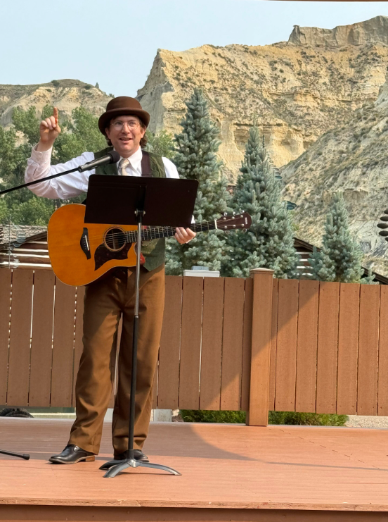 A man wearing a brown hat, white shirt, and vest, playing an acoustic guitar on an outdoor stage with a mountain backdrop and evergreen trees.