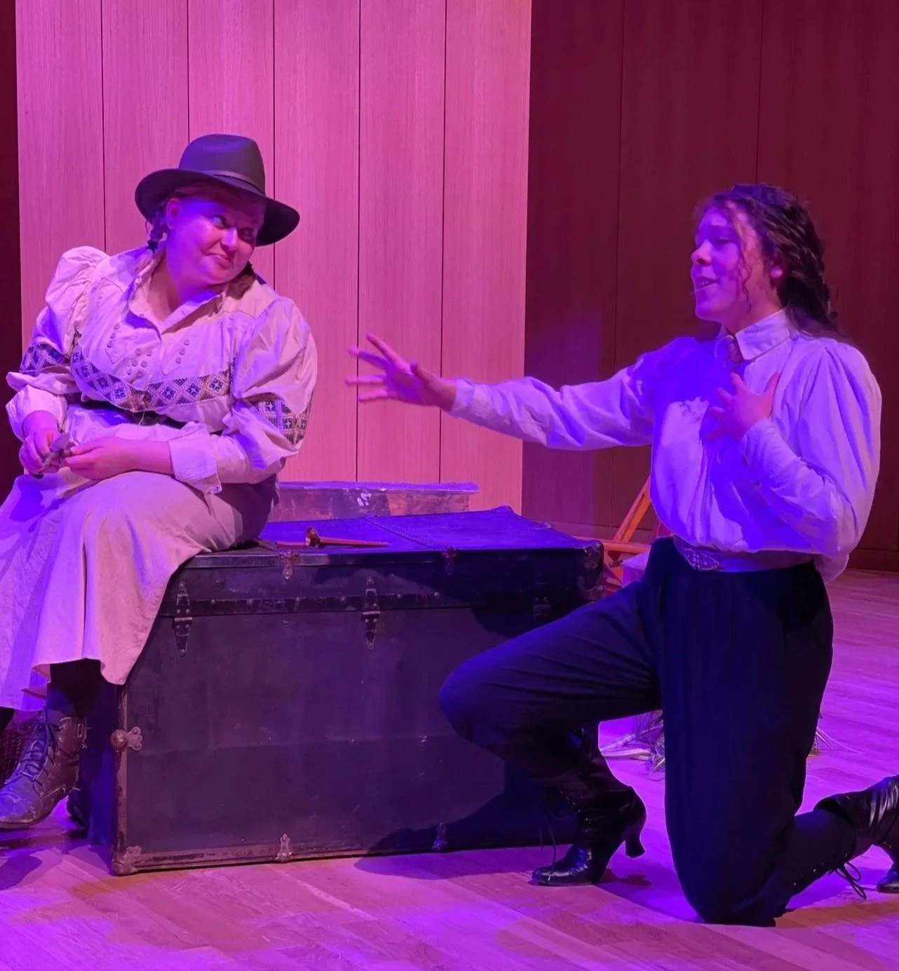 Two women in vintage costumes performing in a theatre, one sitting on a trunk and the other kneeling, engaging in theatrical dialogue.