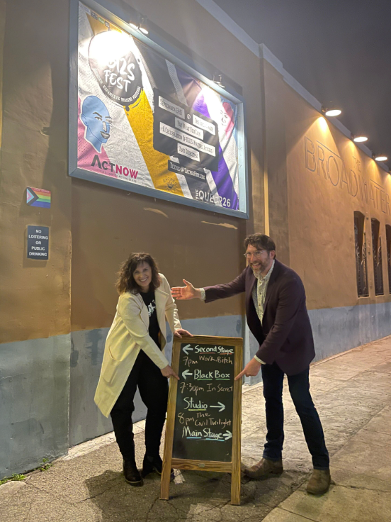 A woman and a man standing on a city sidewalk in front of a theater. They are smiling and pointing at a chalkboard sign. The woman is wearing a cream-colored coat, black pants, and black boots. The man is wearing a dark blazer, light shirt, and dark pants. The chalkboard sign lists various theater stages and showtimes. Behind them, a large illuminated poster for a festival is mounted on the building, and the theater's name 'Broadwater' is on the wall.
