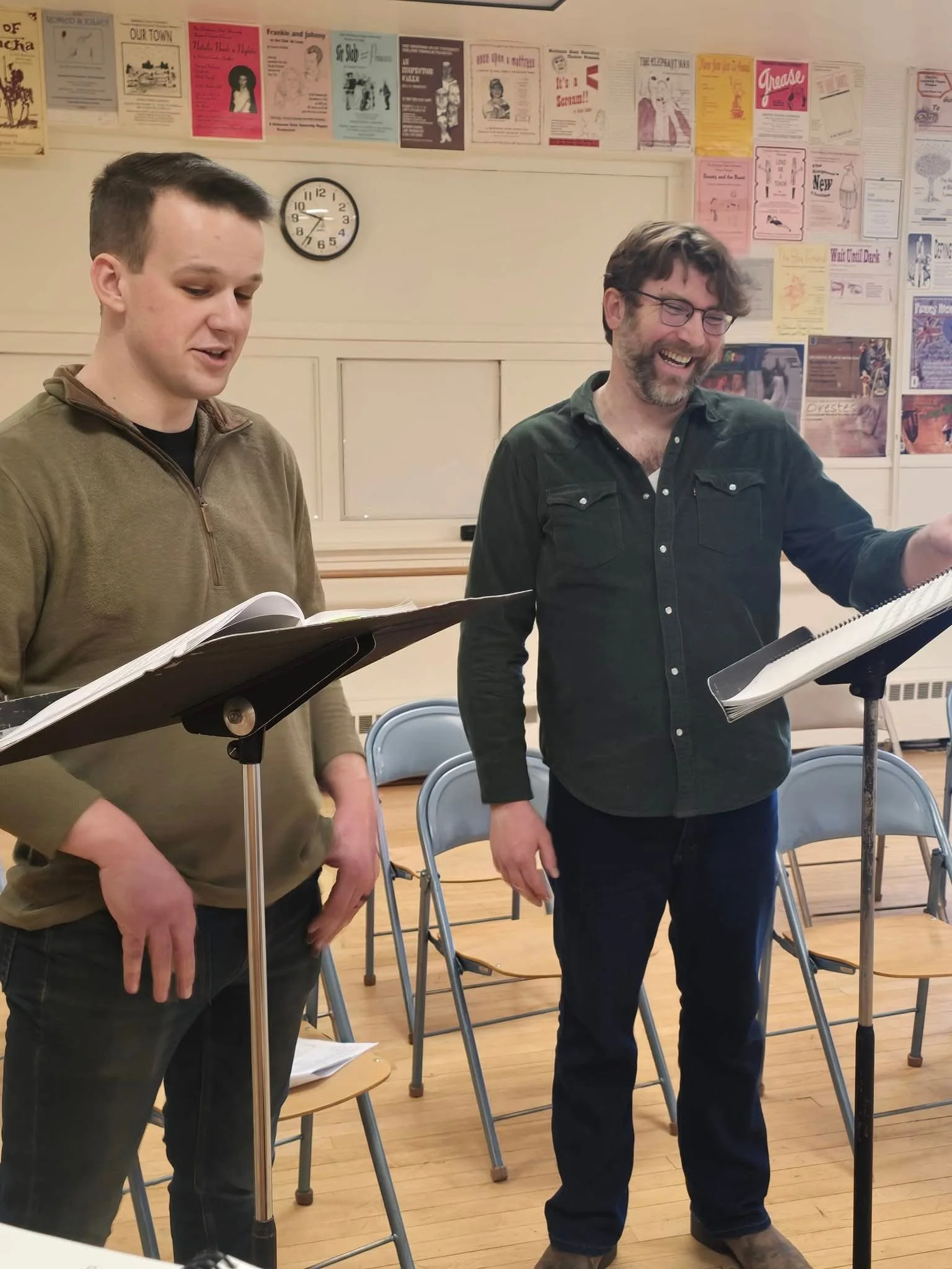 Two men standing in a classroom, reading from scripts on music stands, smiling and laughing; classroom walls decorated with colorful posters and a clock showing 8:11.
