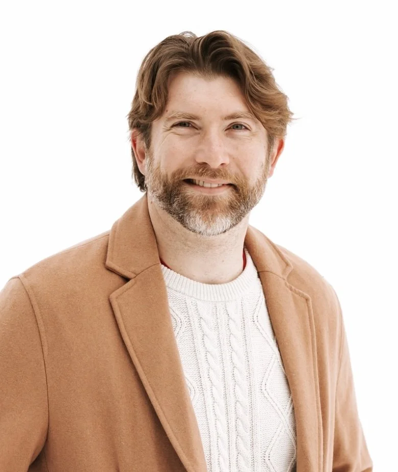 Portrait of a middle-aged man with brown hair and a beard, wearing a tan coat over a white knit sweater, smiling against a white background.