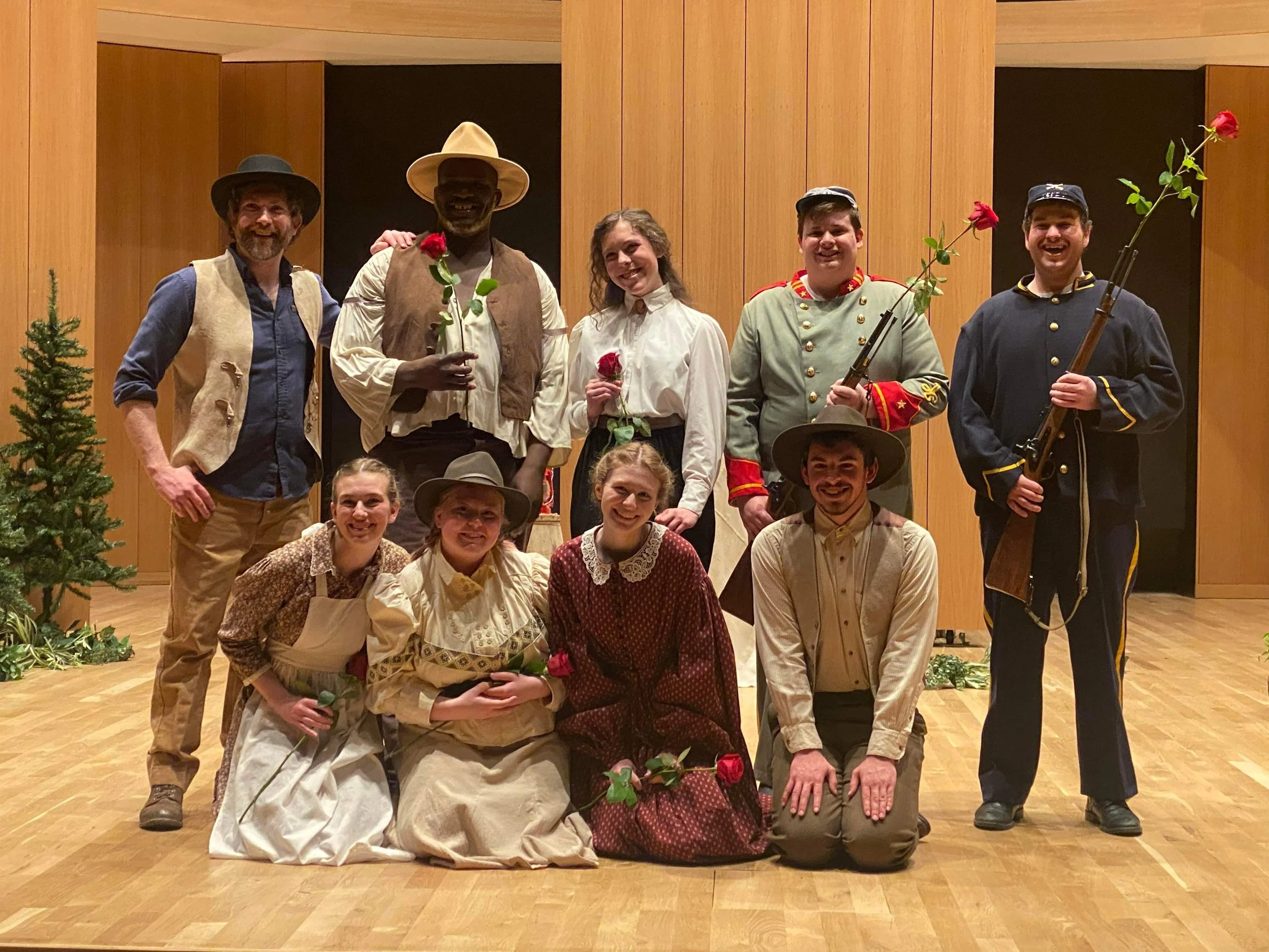 Group of ten people dressed in historical or theatrical costumes, some holding roses and rifles, on a wooden stage with a wood-paneled background, and a small Christmas tree on the left.