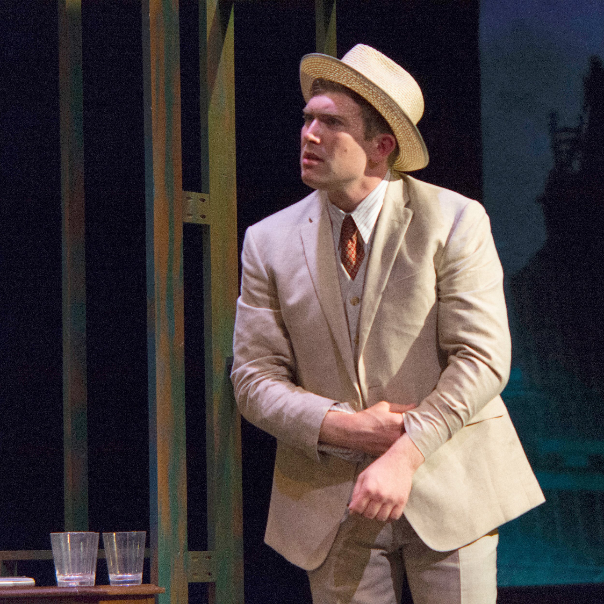 A man wearing a light-colored suit, a straw hat, and a patterned tie, standing on stage with a dark background and some vertical green structures, appearing to be acting in a theatrical performance.