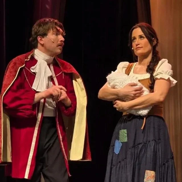 Two actors performing on stage in period costumes, one man with glasses and a red coat, the woman with dark hair in a braid holding a baby doll.