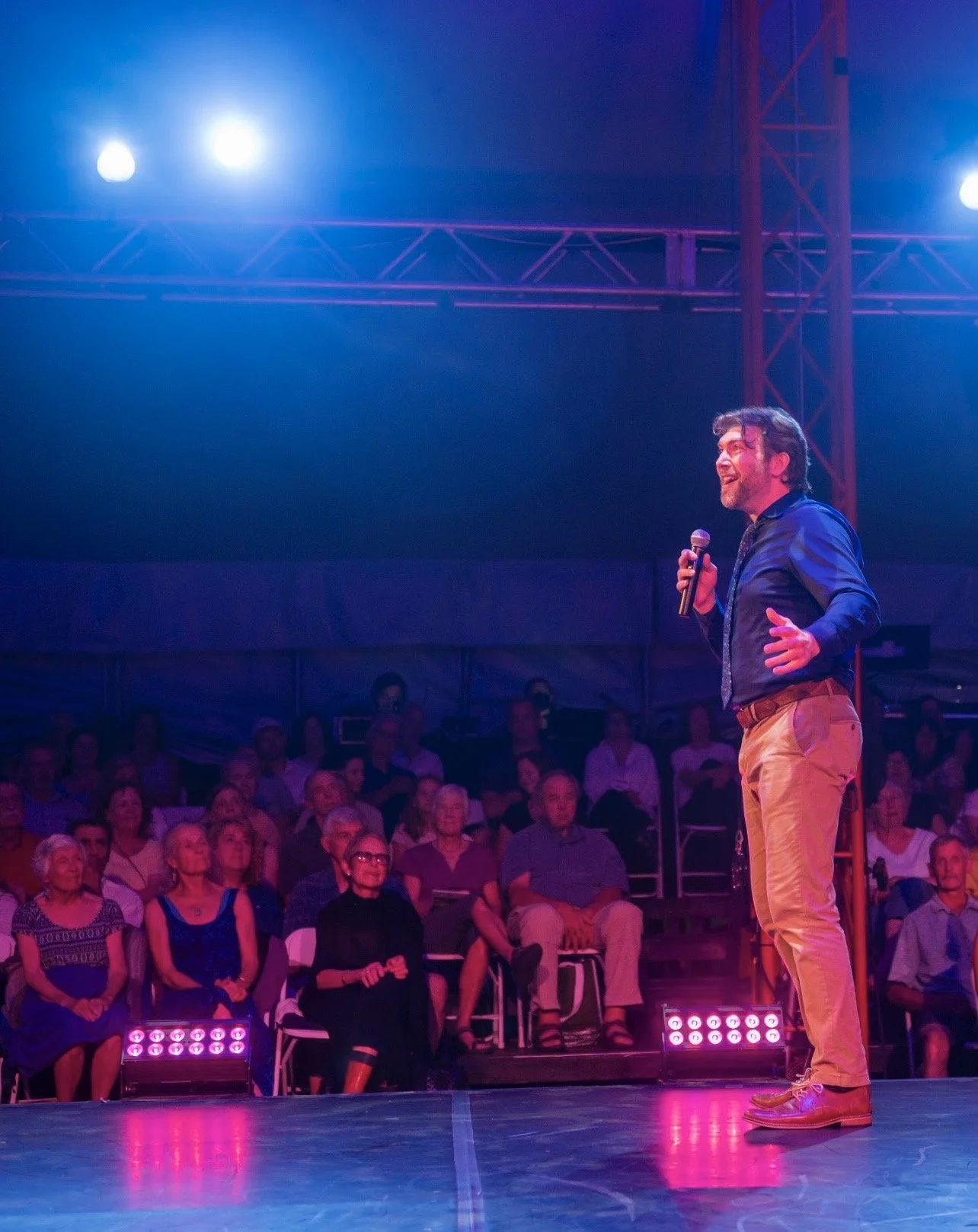 Man on stage speaking into microphone in front of audience at indoor event with stage lighting.
