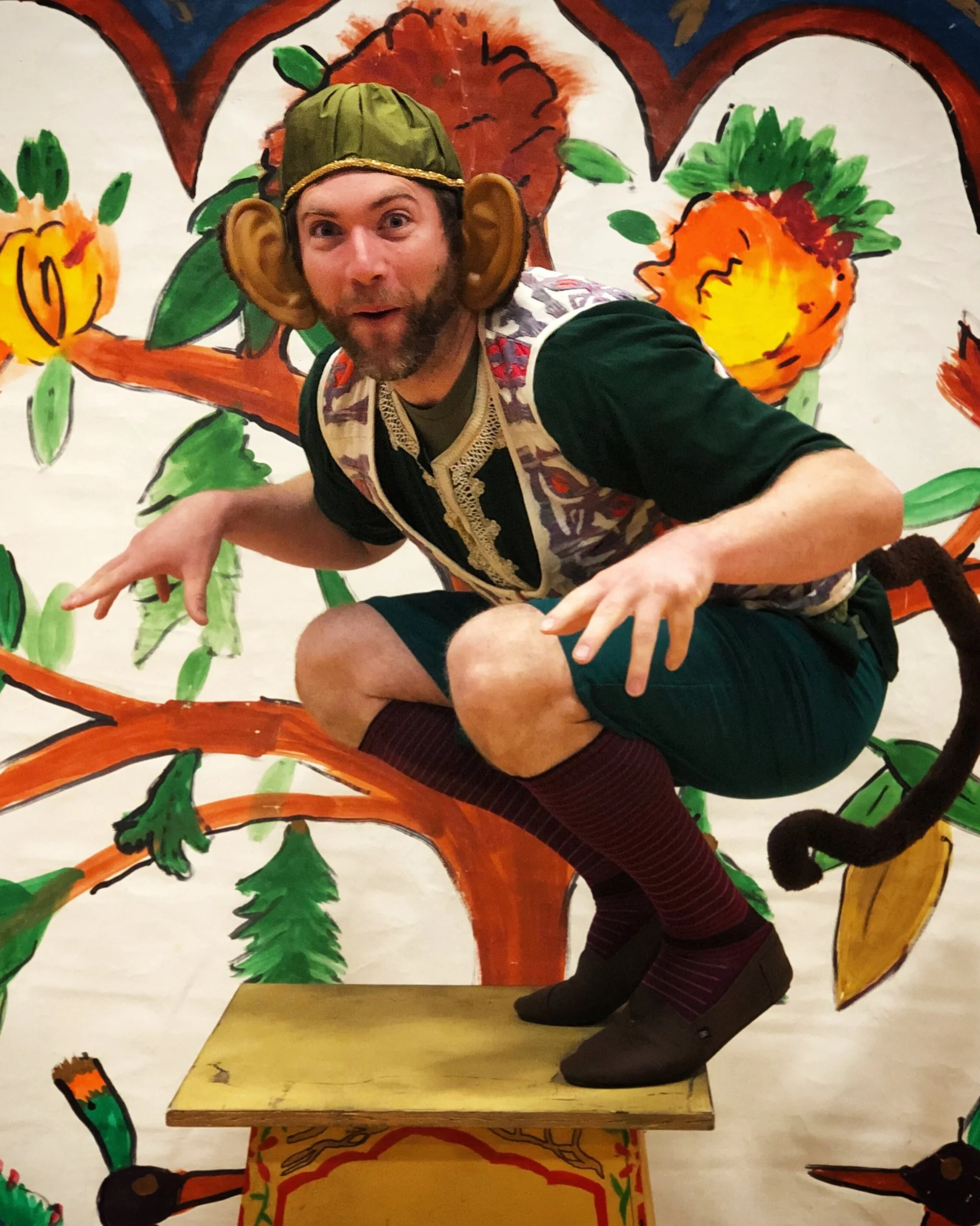 A man dressed in a costume with exaggerated ears, a green hat, and colorful clothing posing on a wooden platform in front of a painted backdrop featuring trees, flowers, and a bear.