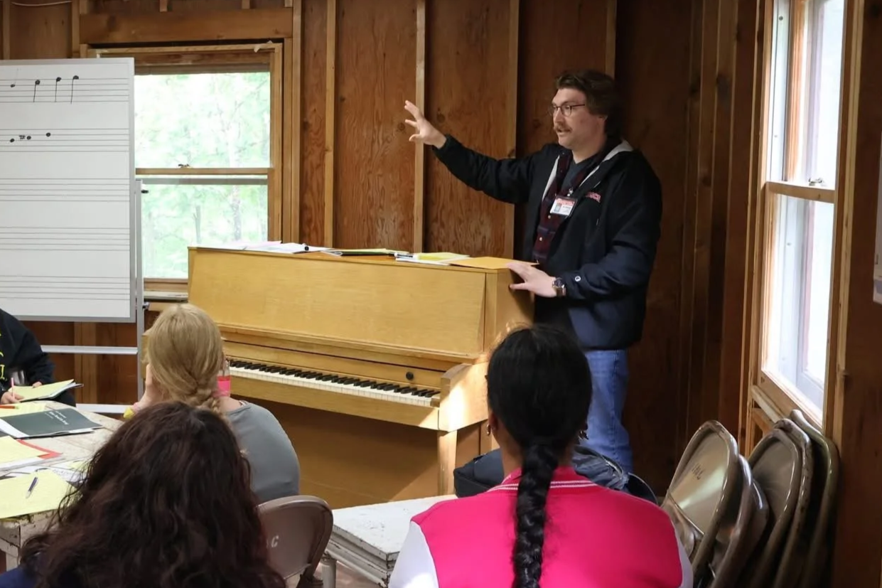 A man teaching a music class, standing near a wooden piano, with students seated around a table, listening attentively in a wood-paneled room with large windows.