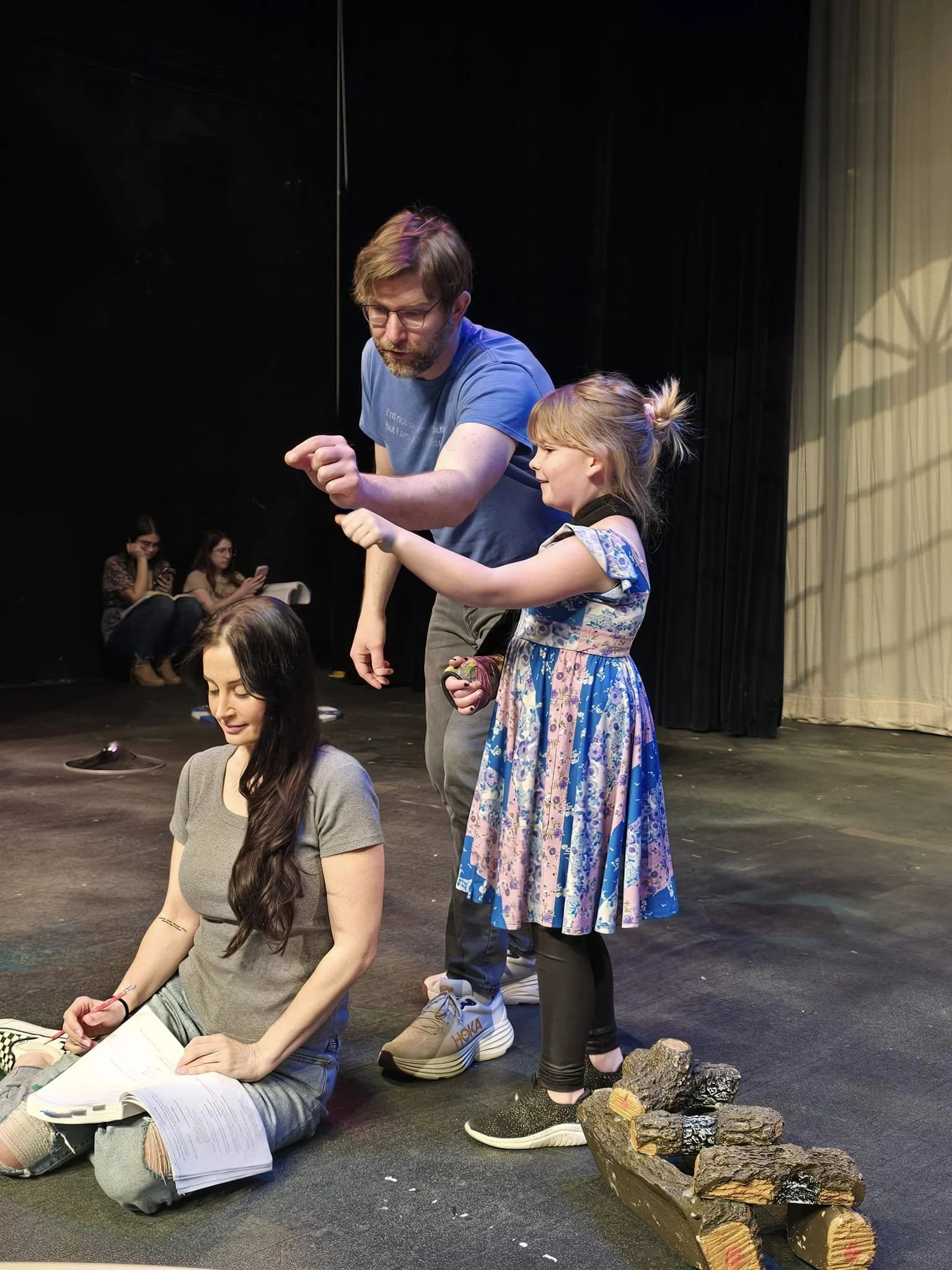 A man and a young girl are rehearsing on a stage; the man shows her a dance move while another woman sits on the floor with a script, taking notes. In the background, two women are sitting and looking at their phones. There is a pile of foil-printed logs on the stage.