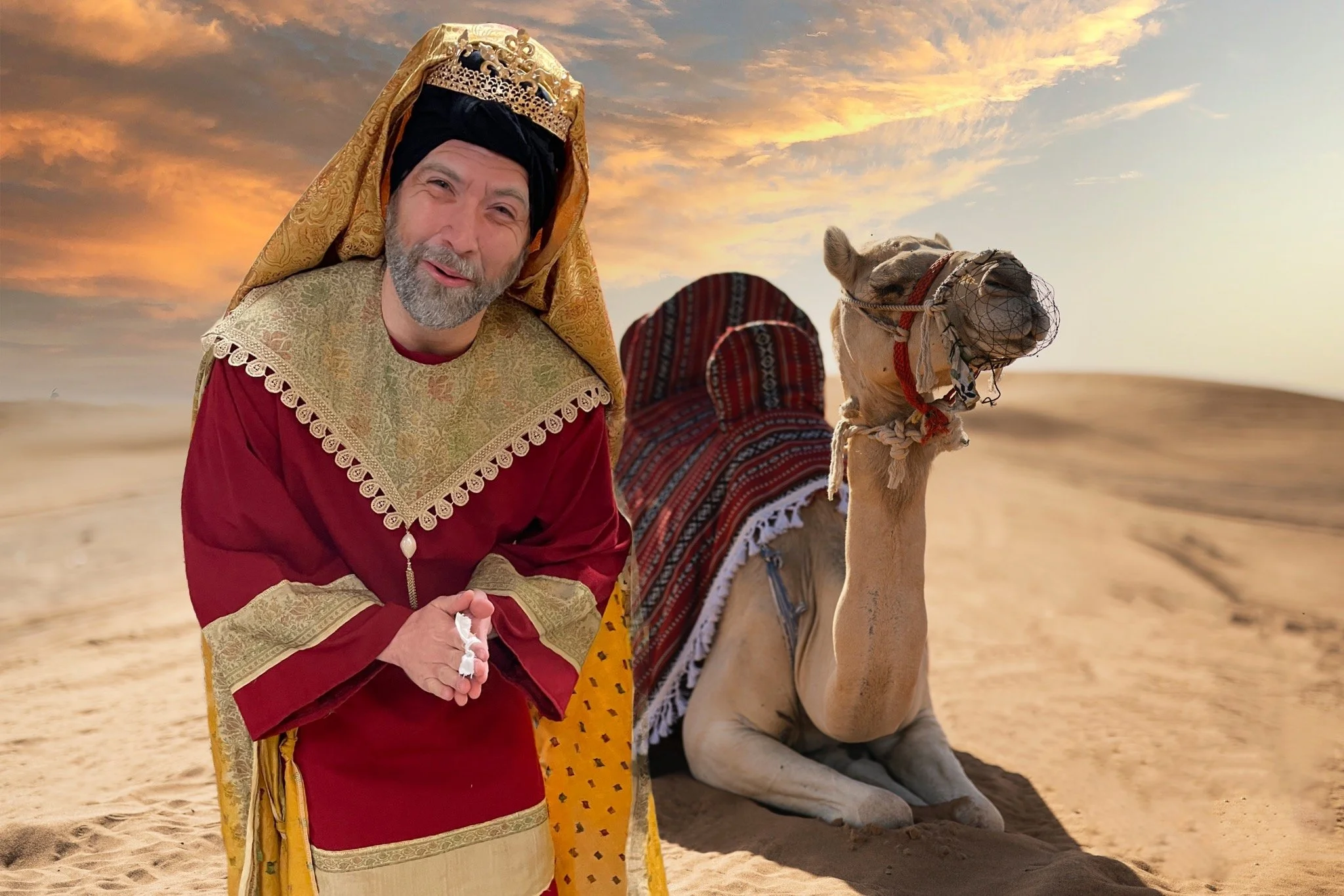A man dressed as a king with a gold crown and royal robes stands in a desert, next to a camel resting on the sand at sunset.