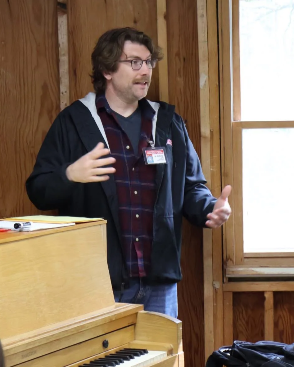 A man with glasses, wearing a black jacket over a plaid shirt, standing indoors near a window and a wooden wall, speaking or explaining something, with a piano in the foreground.