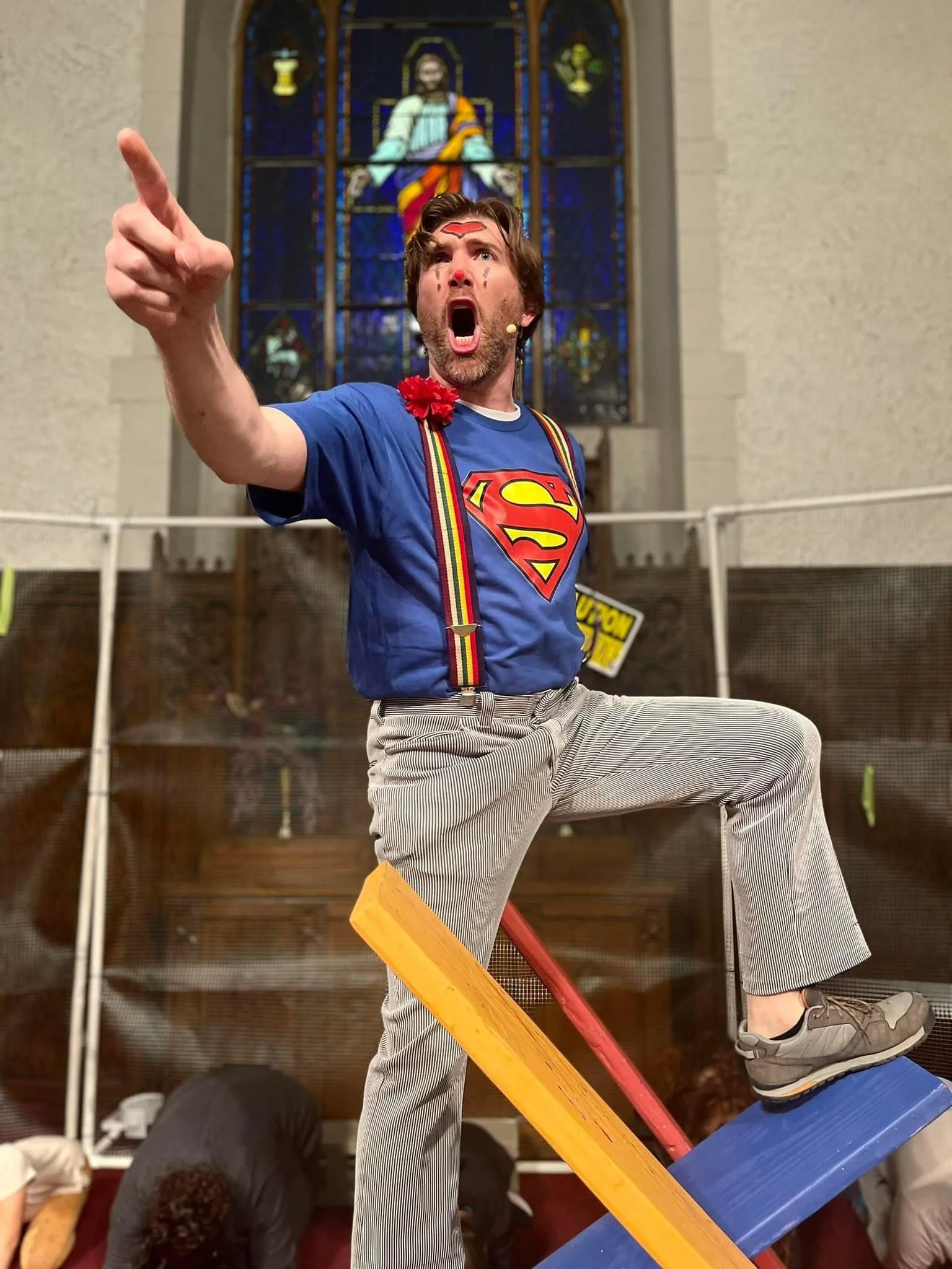 A man dressed as Superman with a red cape, suspenders, and a superhero emblem on his blue shirt is standing on a small platform, reaching forward with an intense expression. He has a painted red mark on his forehead and face, with a stained shirt, in front of a large stained glass window depicting a religious figure.