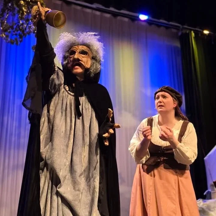 A theatrical stage scene with two actors; one dressed as an old witch with gray curly wig, large mask, and dark cloak, holding a brass bell; the other as a young girl with a headscarf and apron, looking concerned.