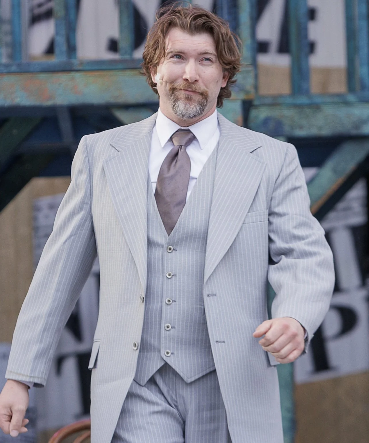 A man with light brown hair, a beard, and mustache wearing a light gray pinstripe suit, white shirt, and brown tie, walking outdoors.