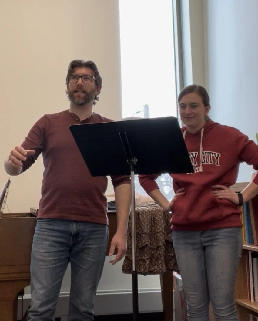 A man and a woman stand near a music stand, inside a room with a large window. The man is wearing glasses and a maroon shirt, the woman is wearing a maroon hoodie with white text and blue jeans.