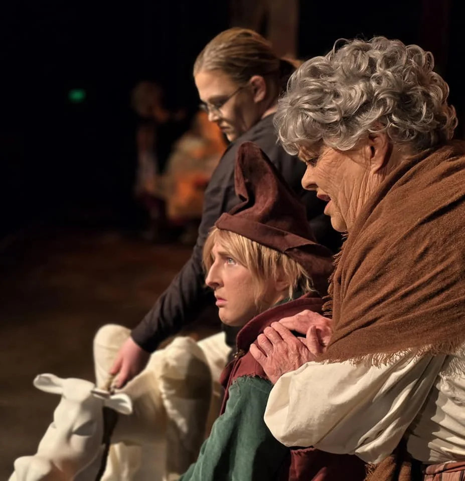 Three women wearing costumes sit in a row on a stage, appearing to be in a theatrical performance.