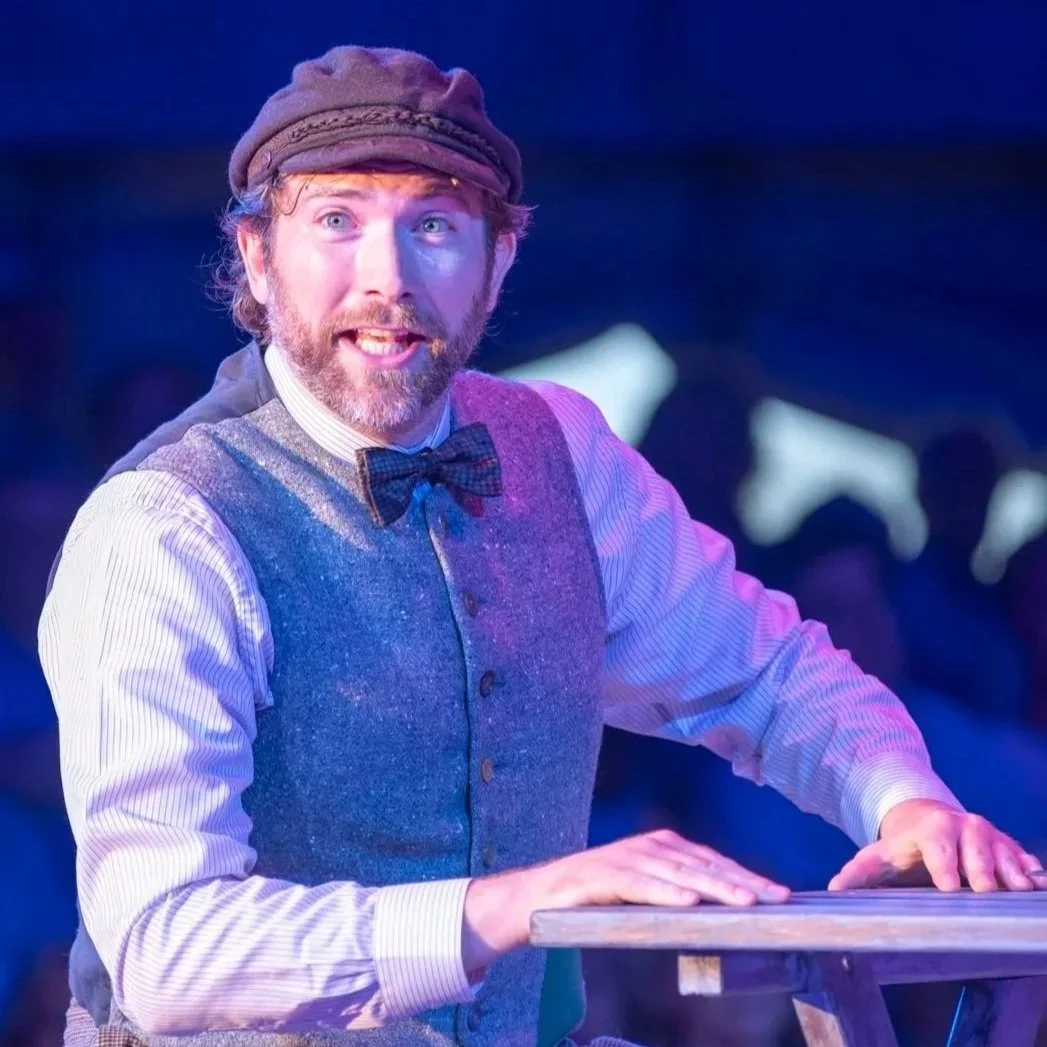 A man with a beard and mustache, wearing a brown cap, striped shirt, vest, and bowtie, is smiling and playing a keyboard or piano on stage with colorful lighting.