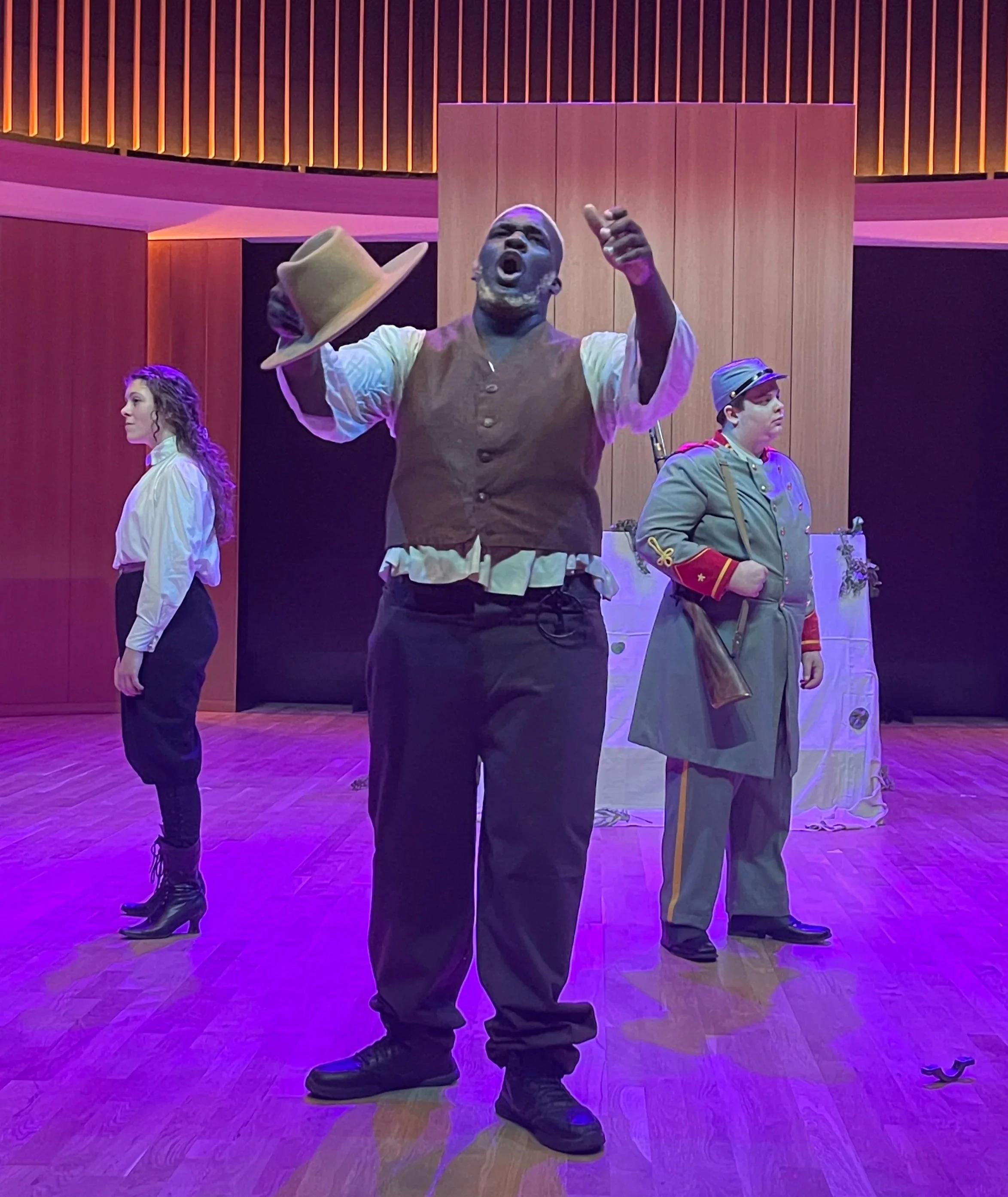 Three actors performing on stage, with one man in the center holding a hat and gesturing, dressed in historical clothing, and two women standing to his sides in period costumes.