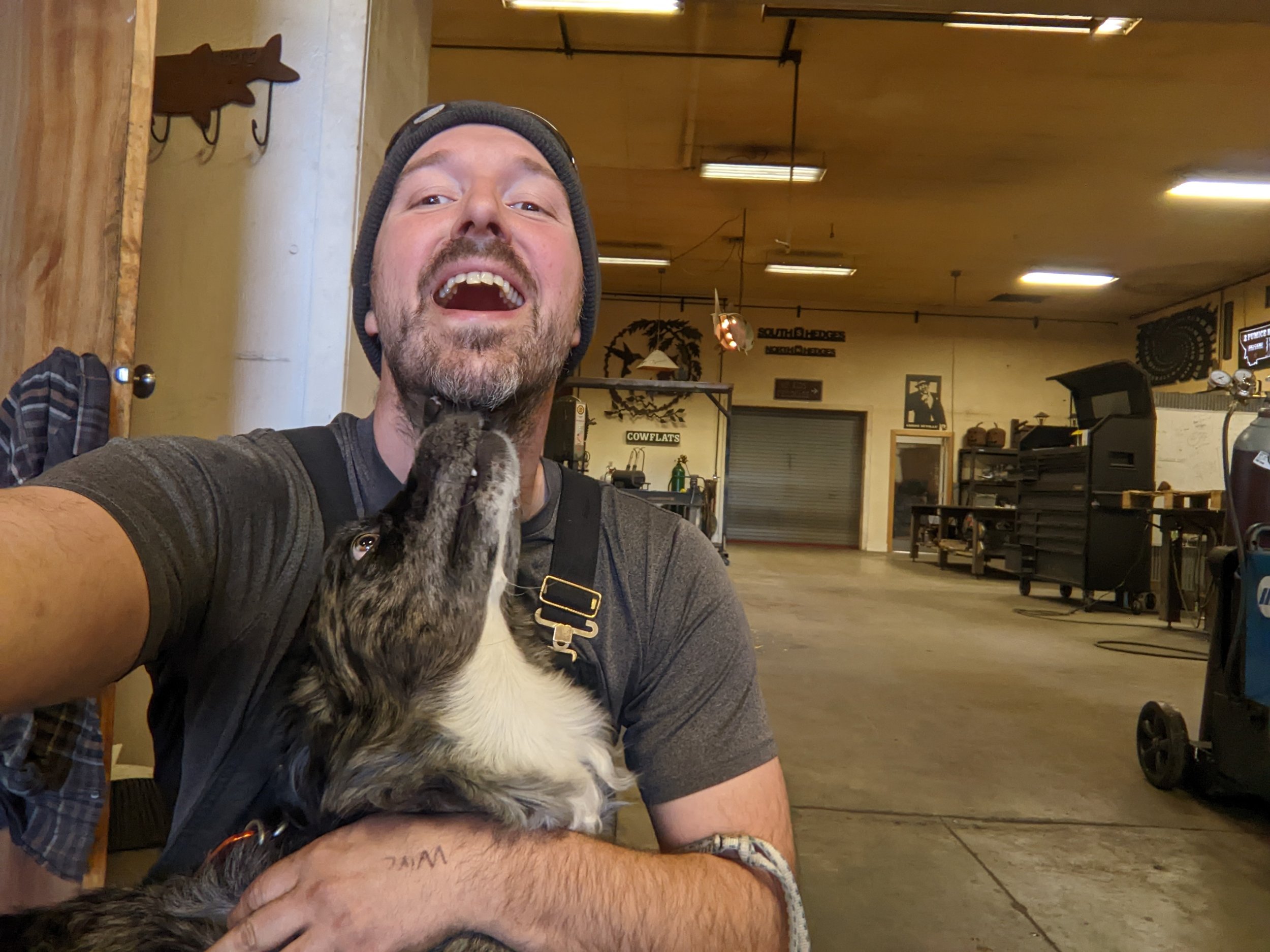 A man with a beard and a beanie, smiling with his mouth open, holding a puppy that is nuzzling his chin, inside a workshop or garage.