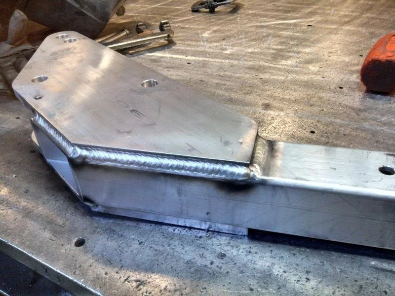 Close-up of a metal component with welded joints, placed on a workbench with tools in the background.