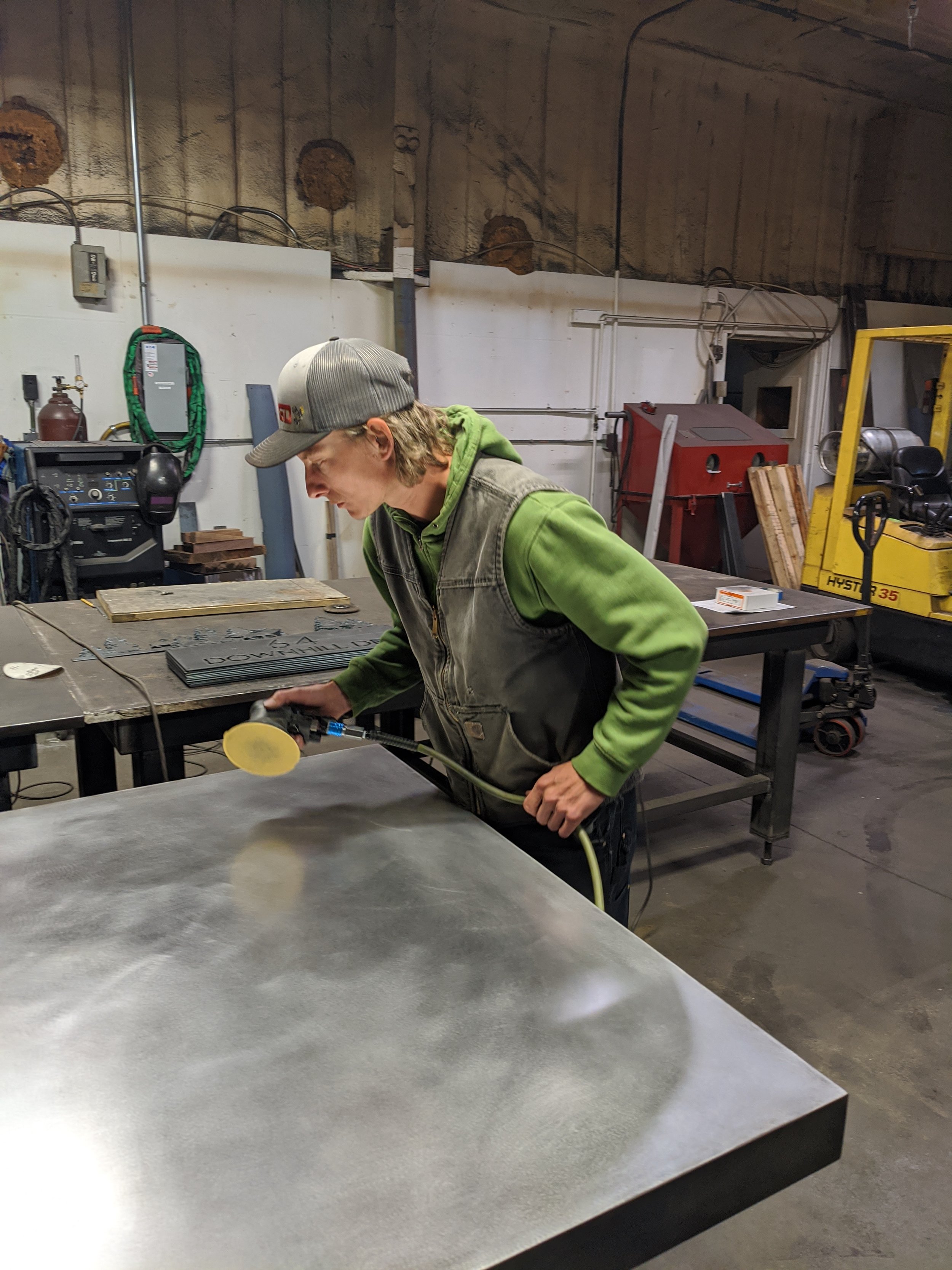 A person wearing a gray cap, green hoodie, and vest is using a pneumatic tool to grind or polish a metal surface in an industrial workshop with various equipment and tools in the background.