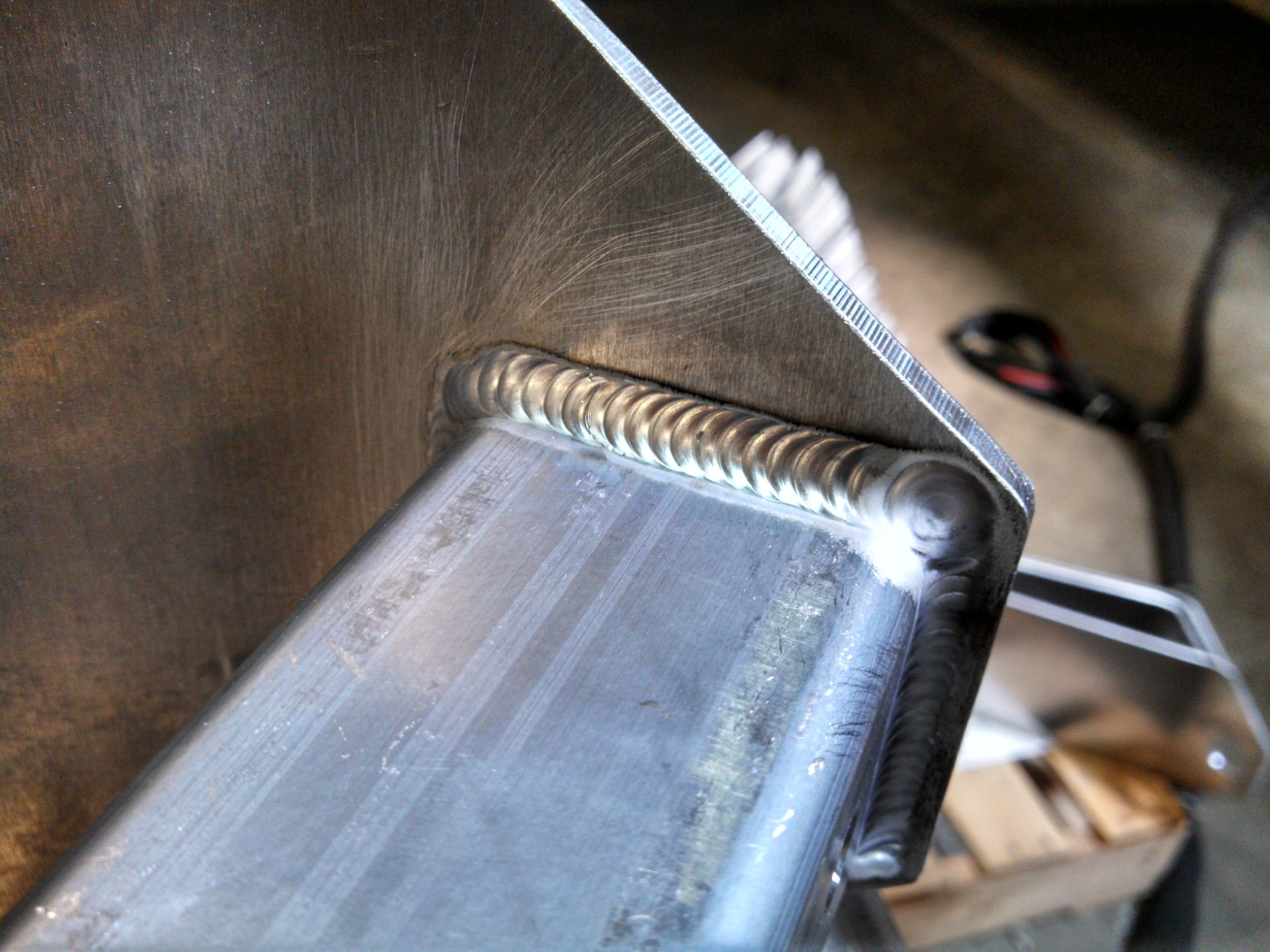 Close-up of a welded metal joint on an aluminum structure, showing a series of weld beads along the corner where two metal pieces meet.