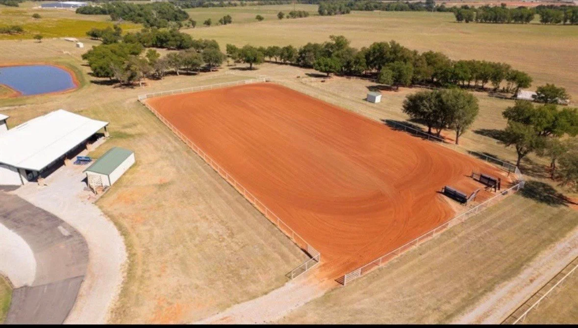 An aerial view of a horse riding arena with reddish dirt, surrounded by trees and fences, with a small building and a pond nearby.