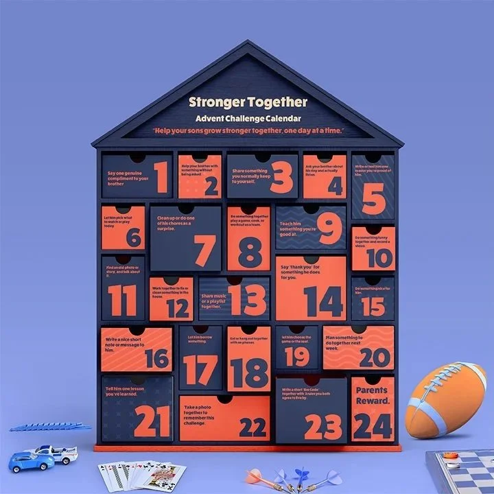 A blue house-shaped advent calendar with 24 numbered compartments, titled 'Stronger Together'. It features tips for parenting and family activities, with some items like toy cars, playing cards, and a miniature football around it.