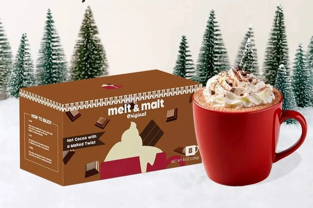 A package of Melt & Mält original hot cocoa with malted twist, a red mug filled with hot chocolate topped with whipped cream and chocolate shavings, set against a snowy background with pine trees.