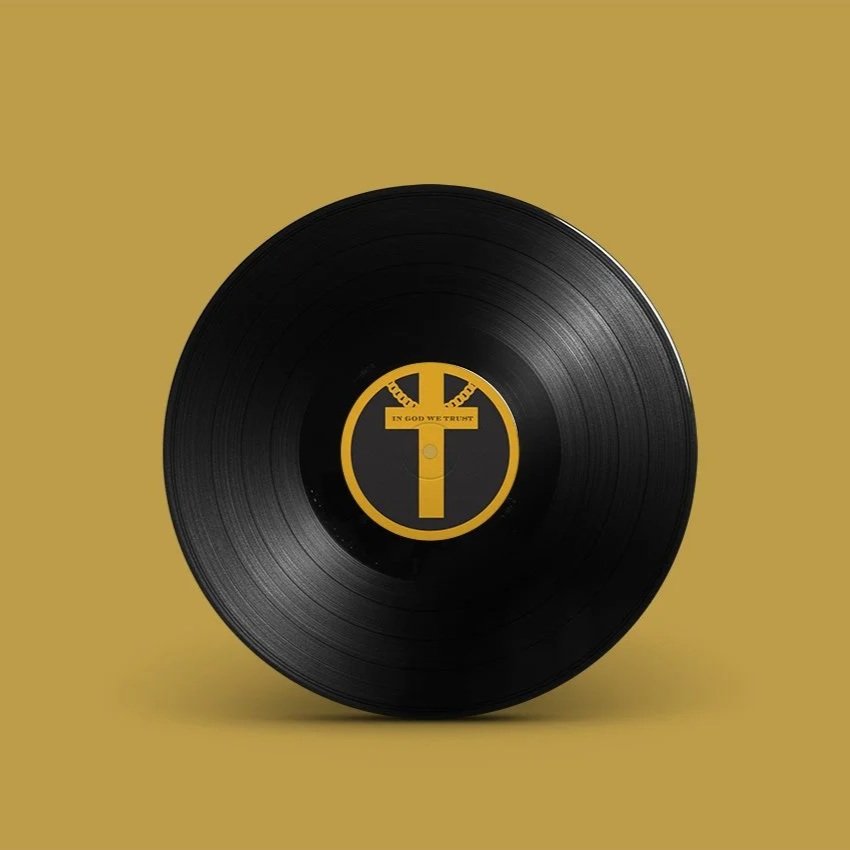 A black vinyl record with a yellow and black label featuring a stylized cross and chain design, with the words "In God We Trust" on a yellow banner.