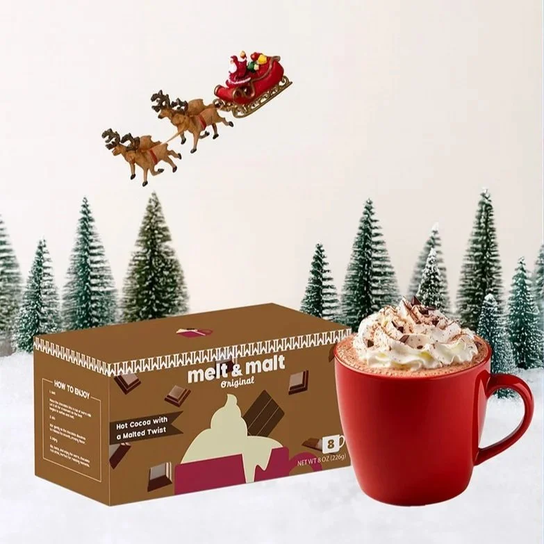 A Christmas-themed scene showing a box of melt and malt hot cocoa, a red mug topped with whipped cream and chocolate shavings, a Santa sleigh with reindeer flying in the sky, and snow-covered pine trees in the background.