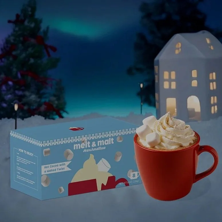 A red mug filled with hot chocolate topped with whipped cream and marshmallows, in front of a winter scene with a Christmas tree, a house, and a starry night sky.