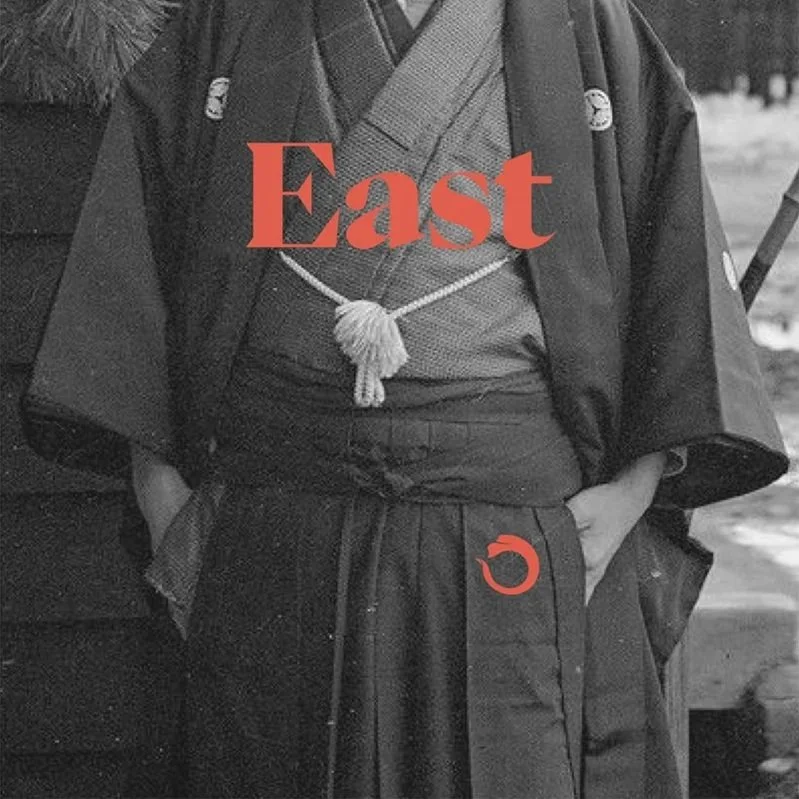 A person dressed in traditional Japanese attire, including a kimono and hakama, standing outdoors with their hands in their pockets. The image has the word 'East' overlaid prominently in red.