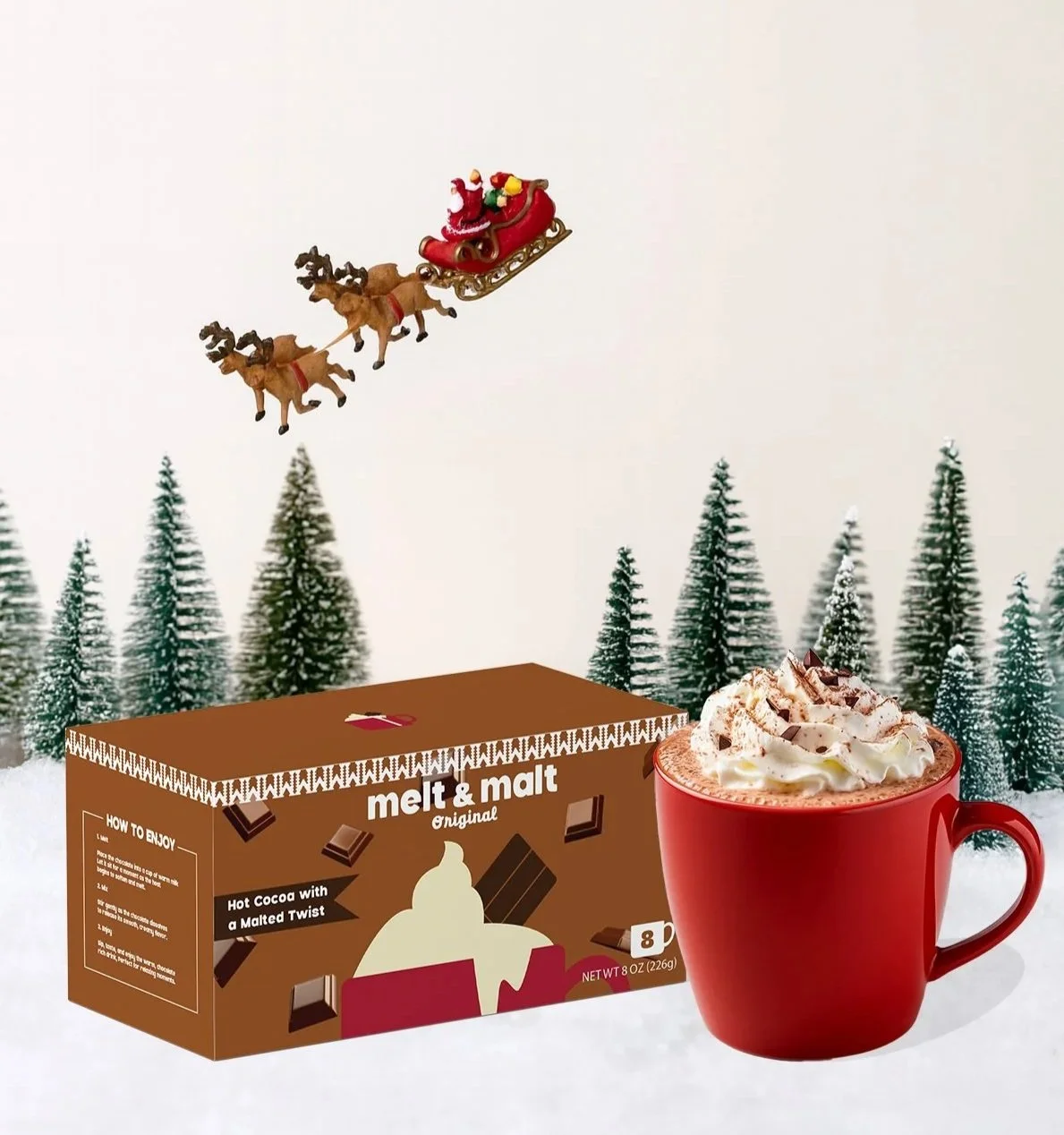 A box of melt and malt hot cocoa mix, a red mug filled with hot cocoa topped with whipped cream and chocolate shavings, and a miniature Santa's sleigh being pulled by two reindeer flying over a snowy landscape with pine trees.
