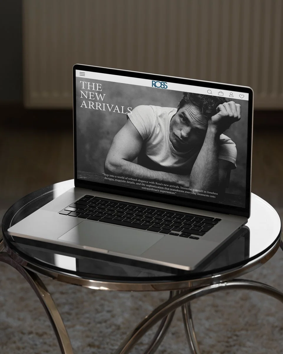 A laptop displaying a fashion website with the headline 'The New Arrivals' and a black-and-white photo of a man with a distressed expression, on a round glass table.