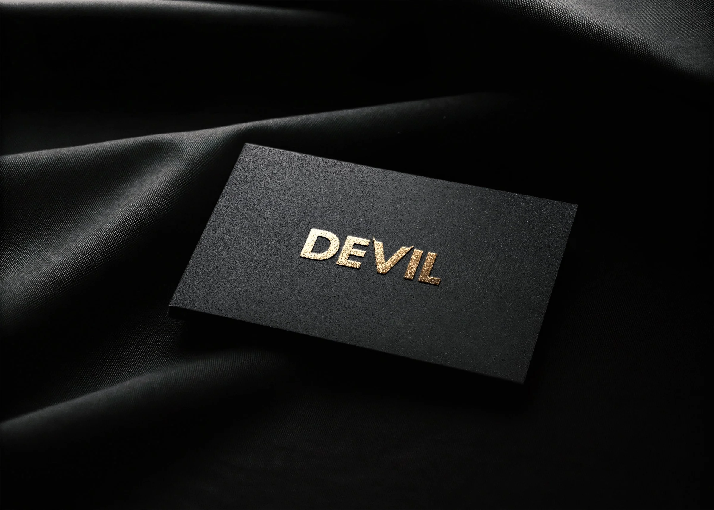 A black textured card with the word 'DEVIL' embossed in metallic gold letters, placed on a black satin fabric.