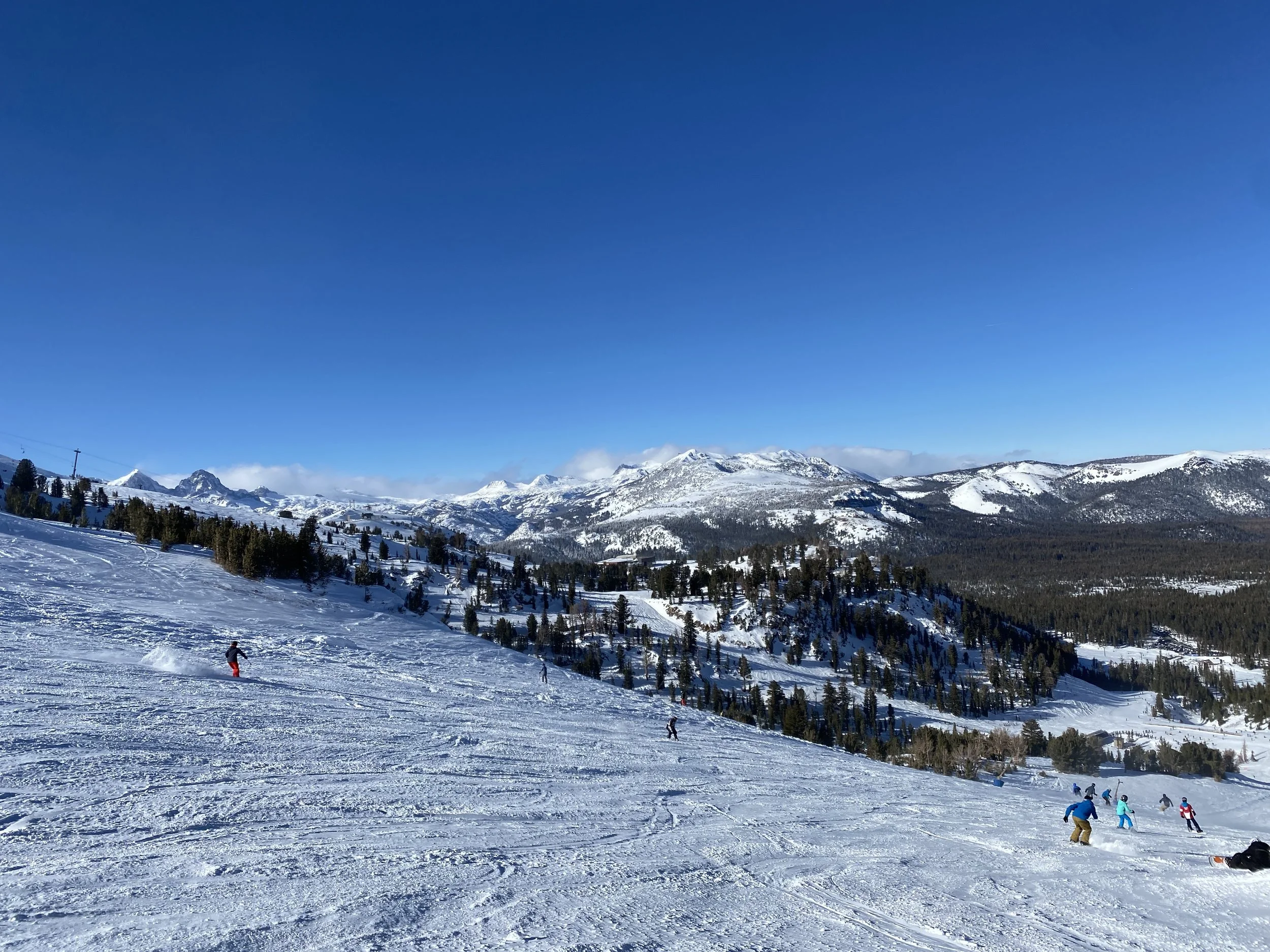 Snow-covered mountain landscape with skiers and snowboarders on the slope under a clear blue sky.