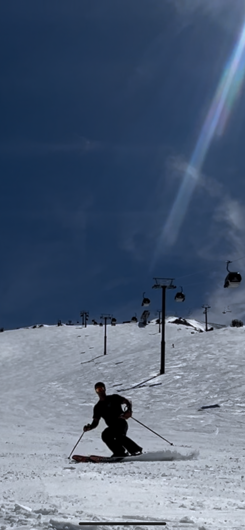 Skier on snowy slope under clear blue sky with ski lifts and a sun flare.