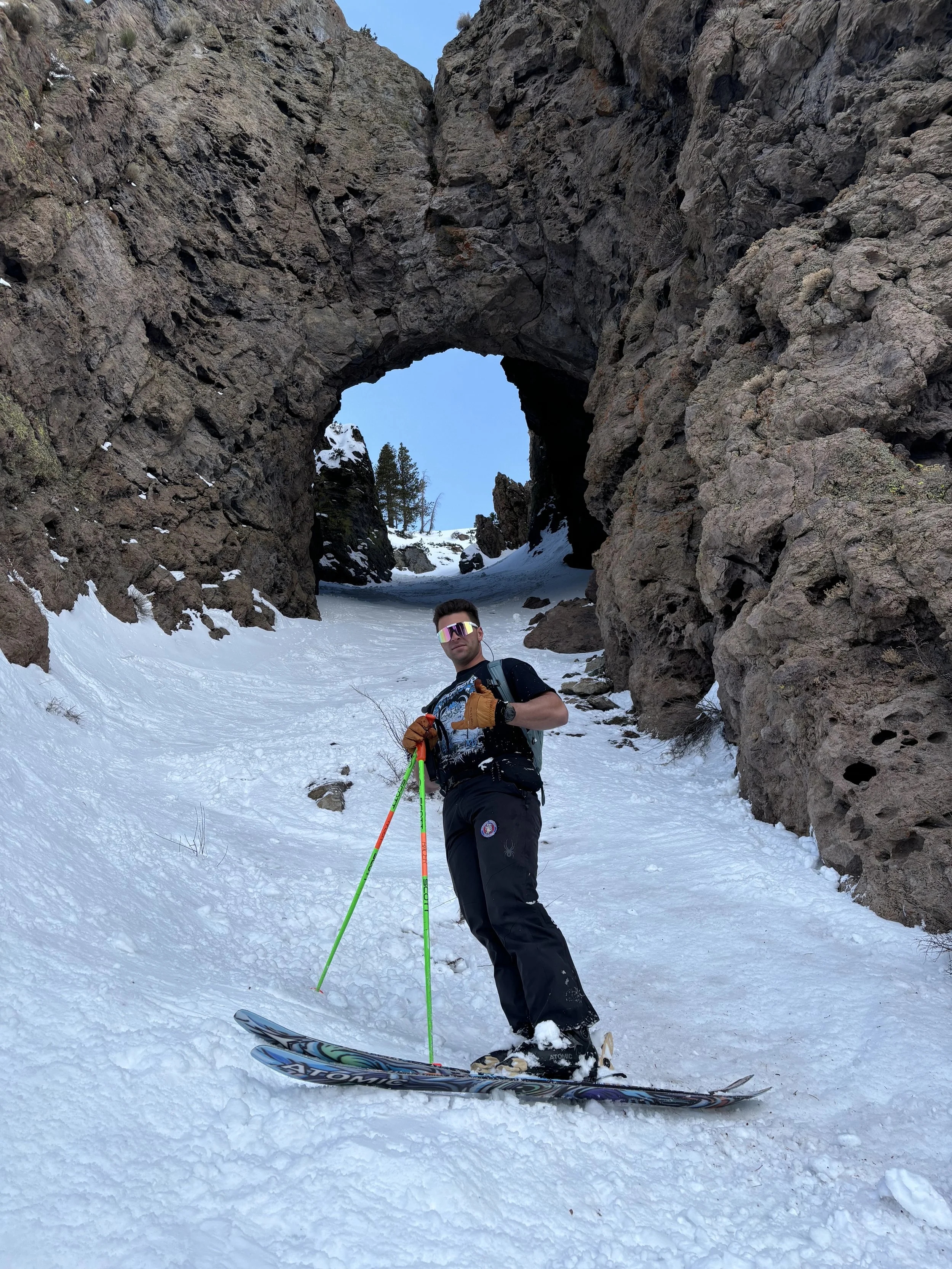 A man wearing ski gear, sunglasses, and a black backpack stands on snow while holding ski poles in a snowy mountain landscape with a rock arch and cliffs in the background.