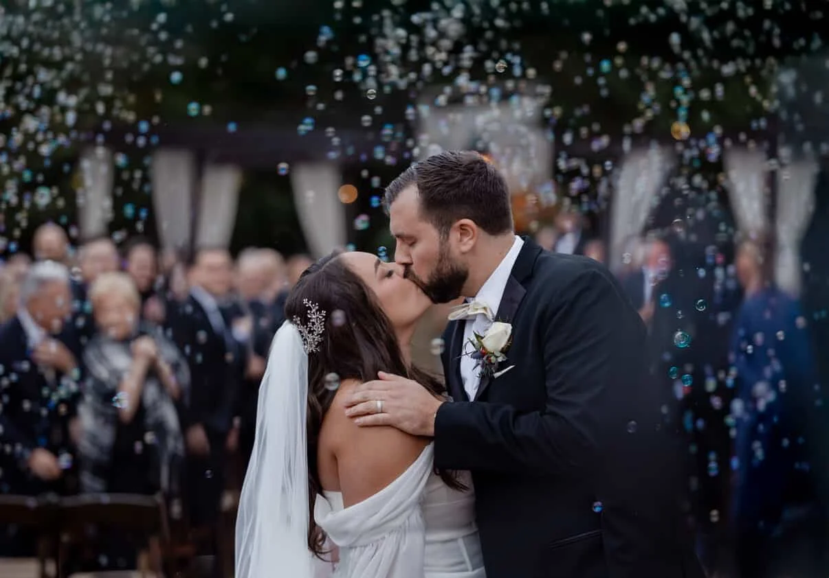 A bride and groom kiss during their wedding ceremony with guests in the background and bubbles floating around them.
