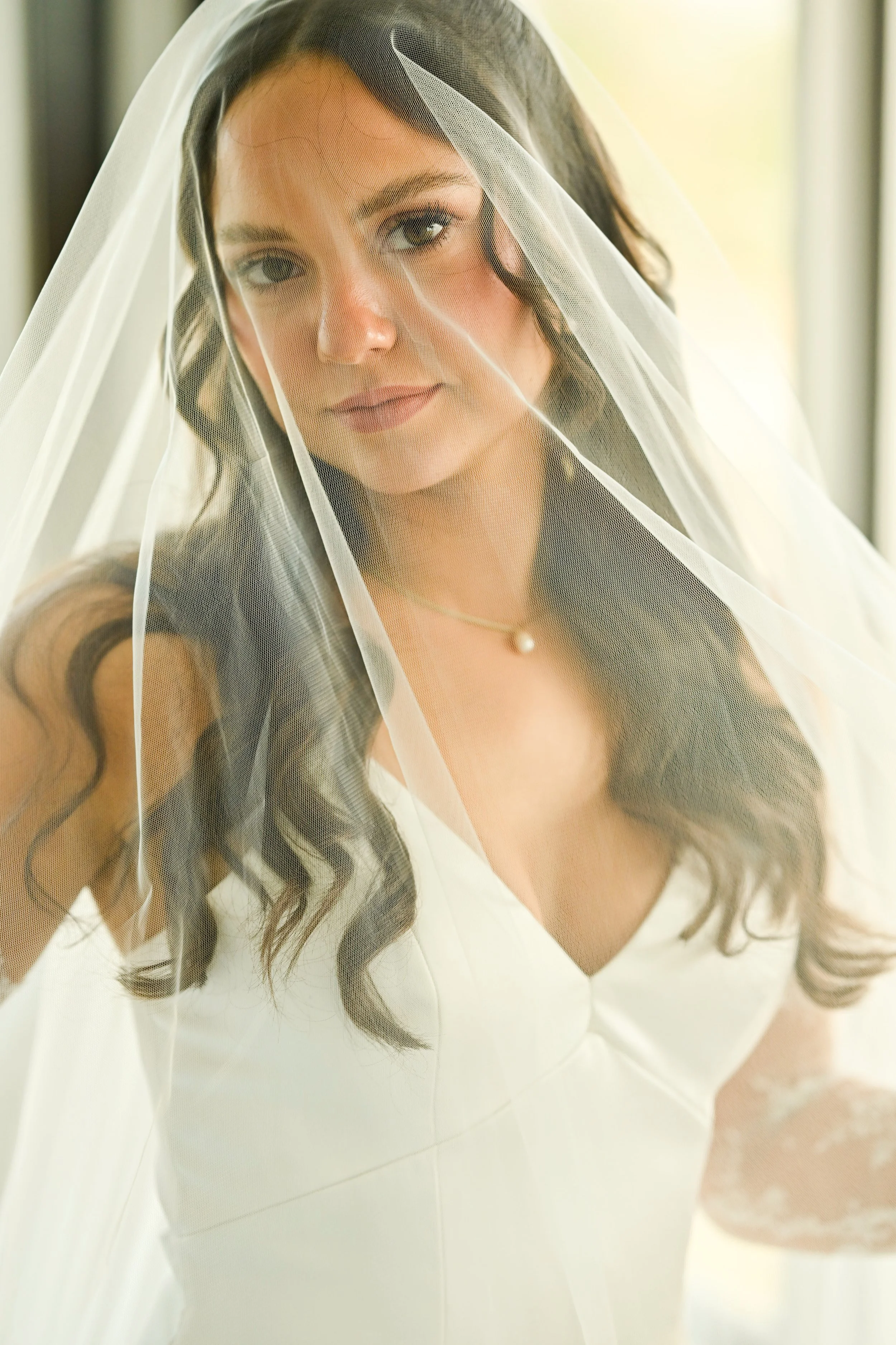 A woman with long dark hair, wearing a white dress and a pearl necklace, is standing behind semi-transparent white curtains, with sunlight coming through a window and illuminating her face.